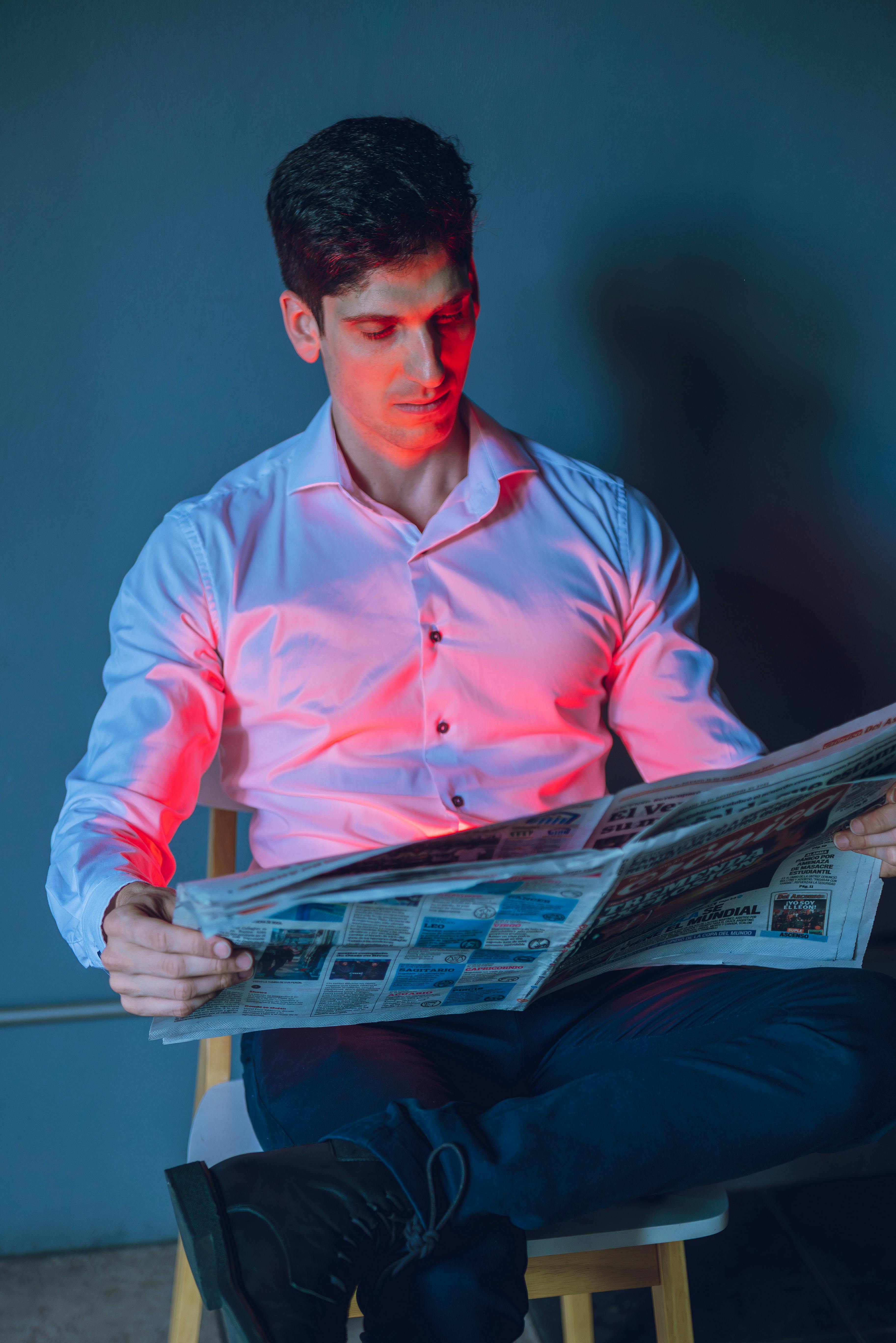 Gratis Un giovane uomo con una camicia bianca legge il giornale sotto una suggestiva illuminazione rossa e blu in un interno. Foto a disposizione