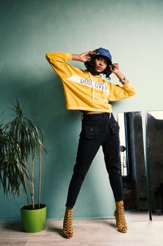 A fashionable woman poses indoors in a trendy yellow hoodie and jeans.