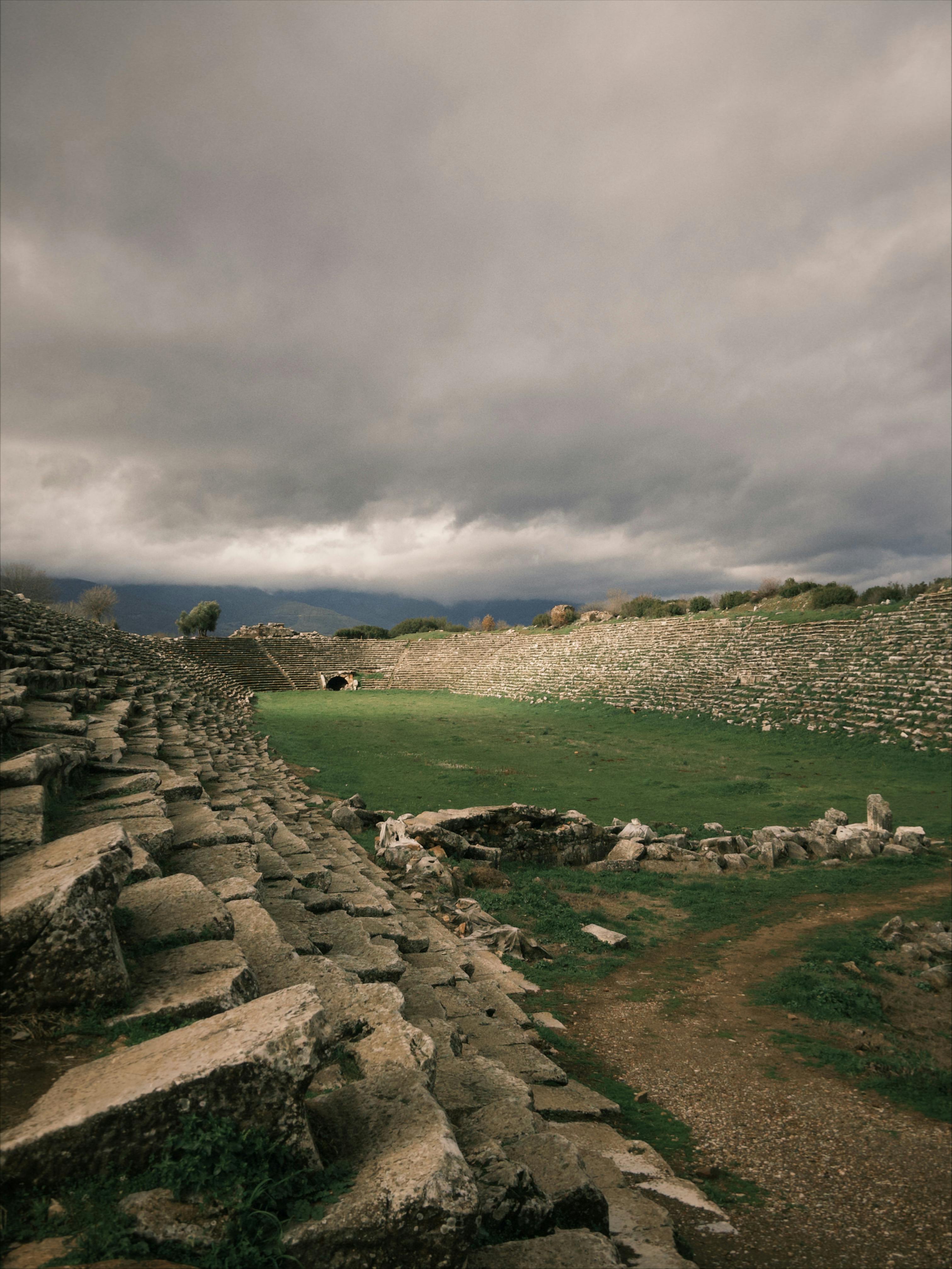 Gratis Temukan amfiteater bersejarah di Aydın, Turki, yang dibingkai oleh awan dramatis dan bangunan batu kuno. Foto Stok