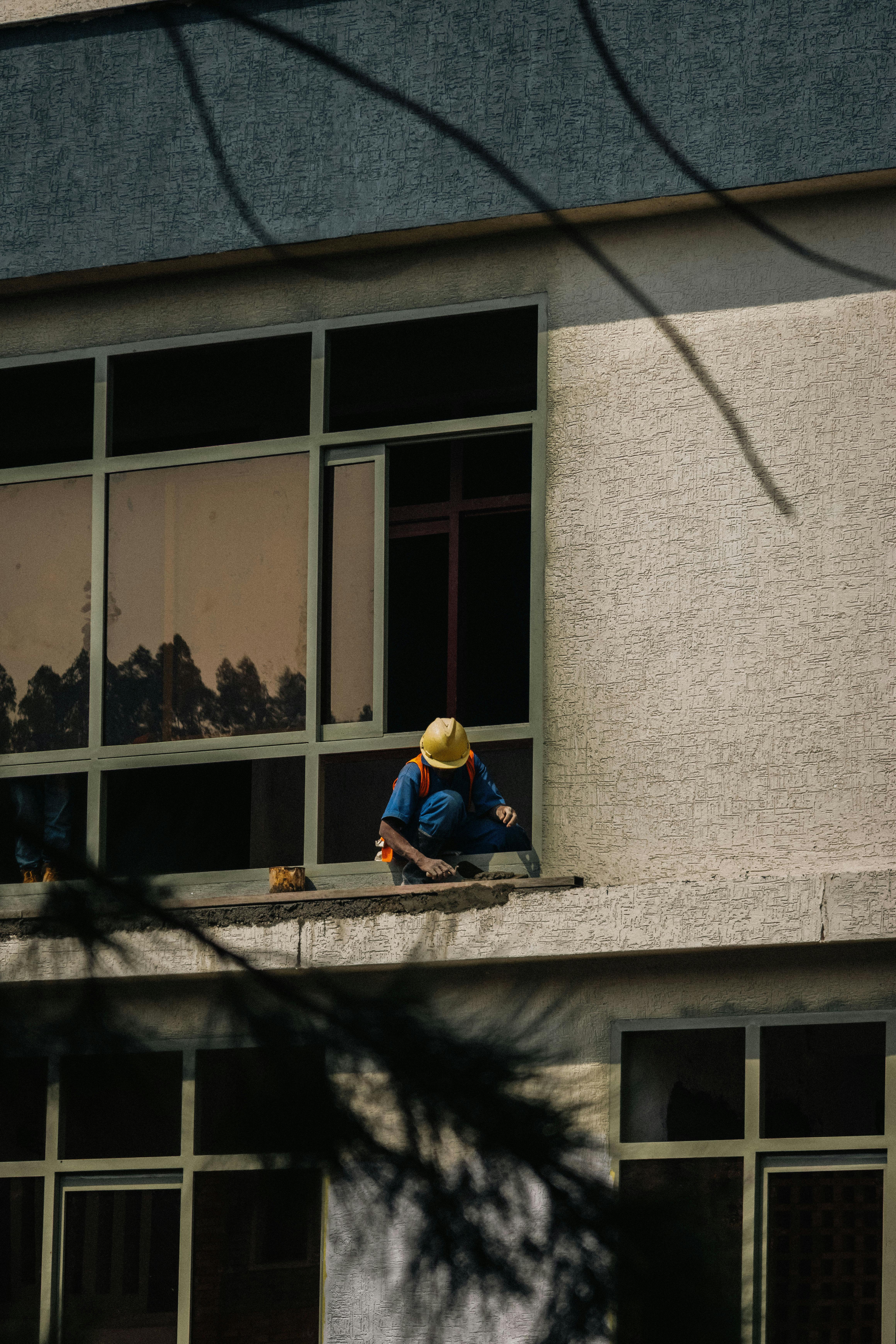 ingyenes Biztonsági felszerelést viselő munkás javítja az épület homlokzatát egy kültéri párkányról. Stockfotó