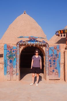Woman standing at a traditional Middle Eastern mud building with blue doors under the clear blue sky.