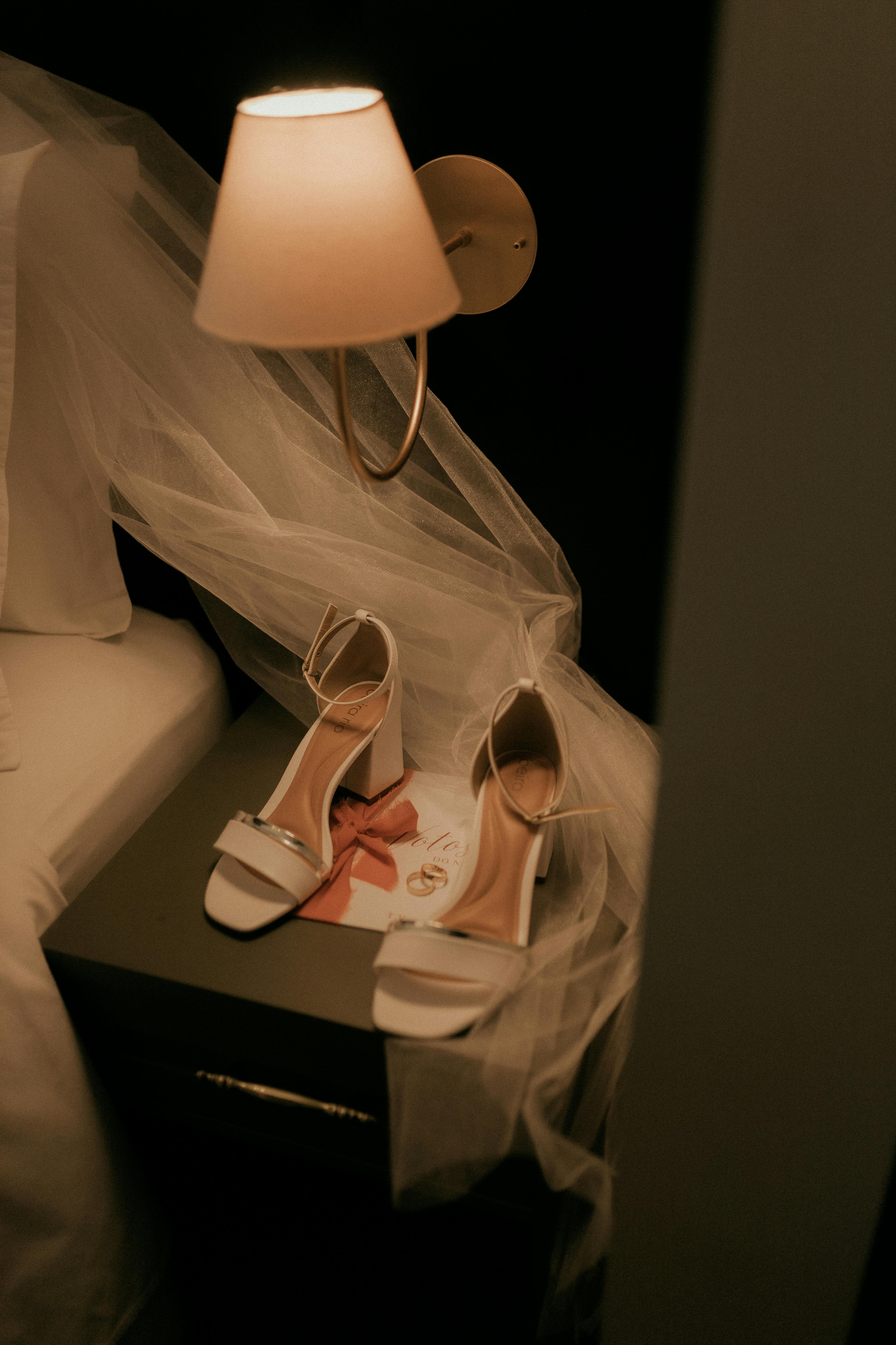 Δωρεάν Ένα ζευγάρι κομψά λευκά νυφικά παπούτσια τοποθετημένα κάτω από ένα απαλό φωτιστικό τοίχου δίπλα σε ένα κρεβάτι, ντυμένο με τούλι. Φωτογραφία από στοκ φωτογραφιών