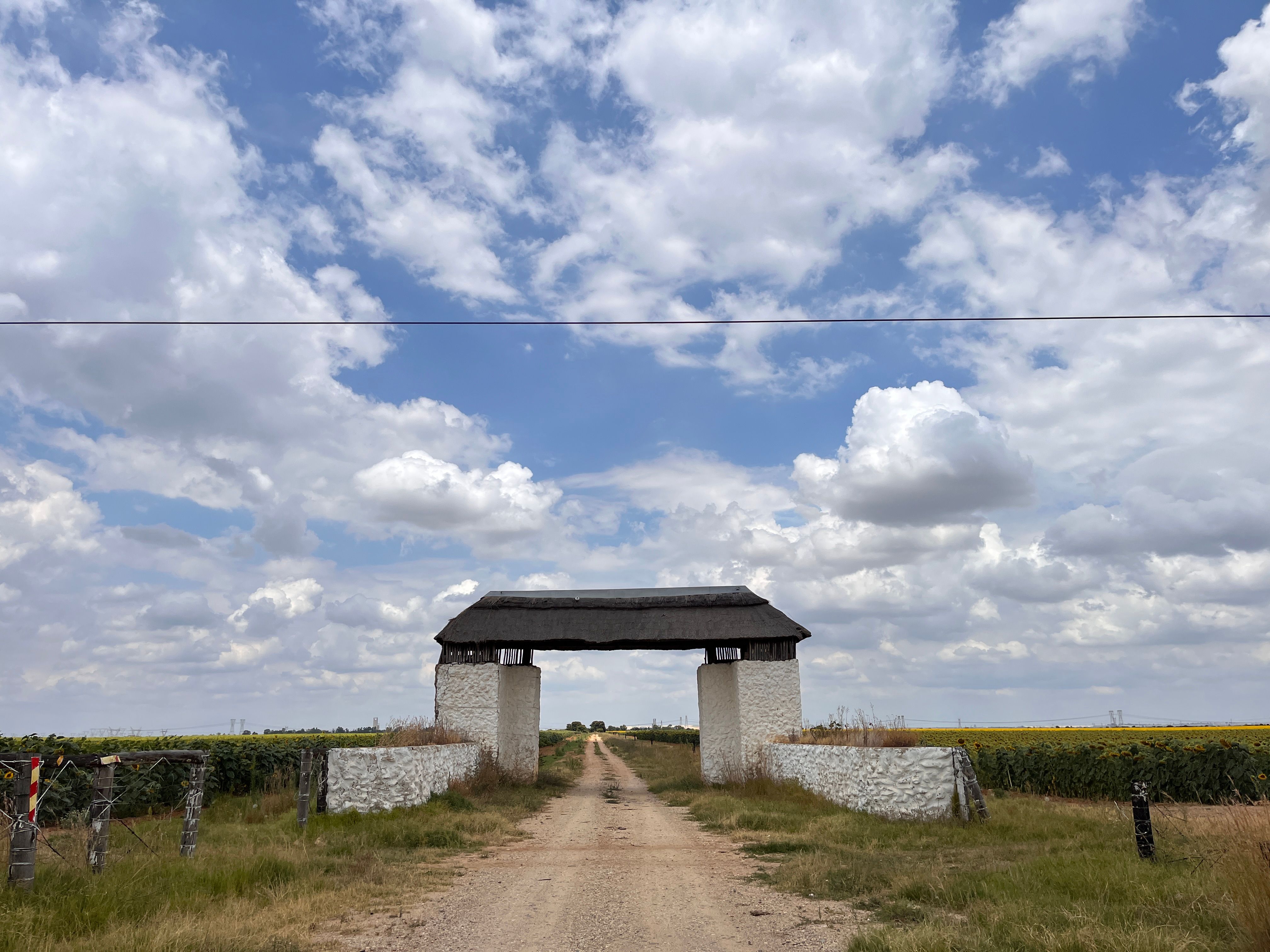 A scenic rural dirt road framed by a rustic entrance against a backdrop of expansive farmland and a vibrant blue sky.