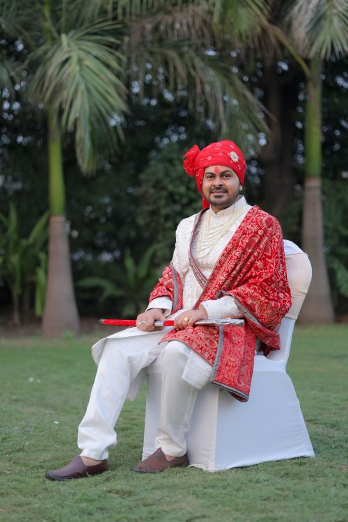 Free Indian groom in vibrant red attire seated outdoors, showcasing traditional wedding style. Stock Photo