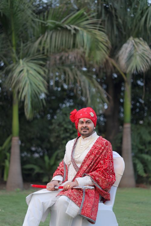 Free A groom in a red turban and traditional attire sits outdoors in a lush garden setting. Stock Photo