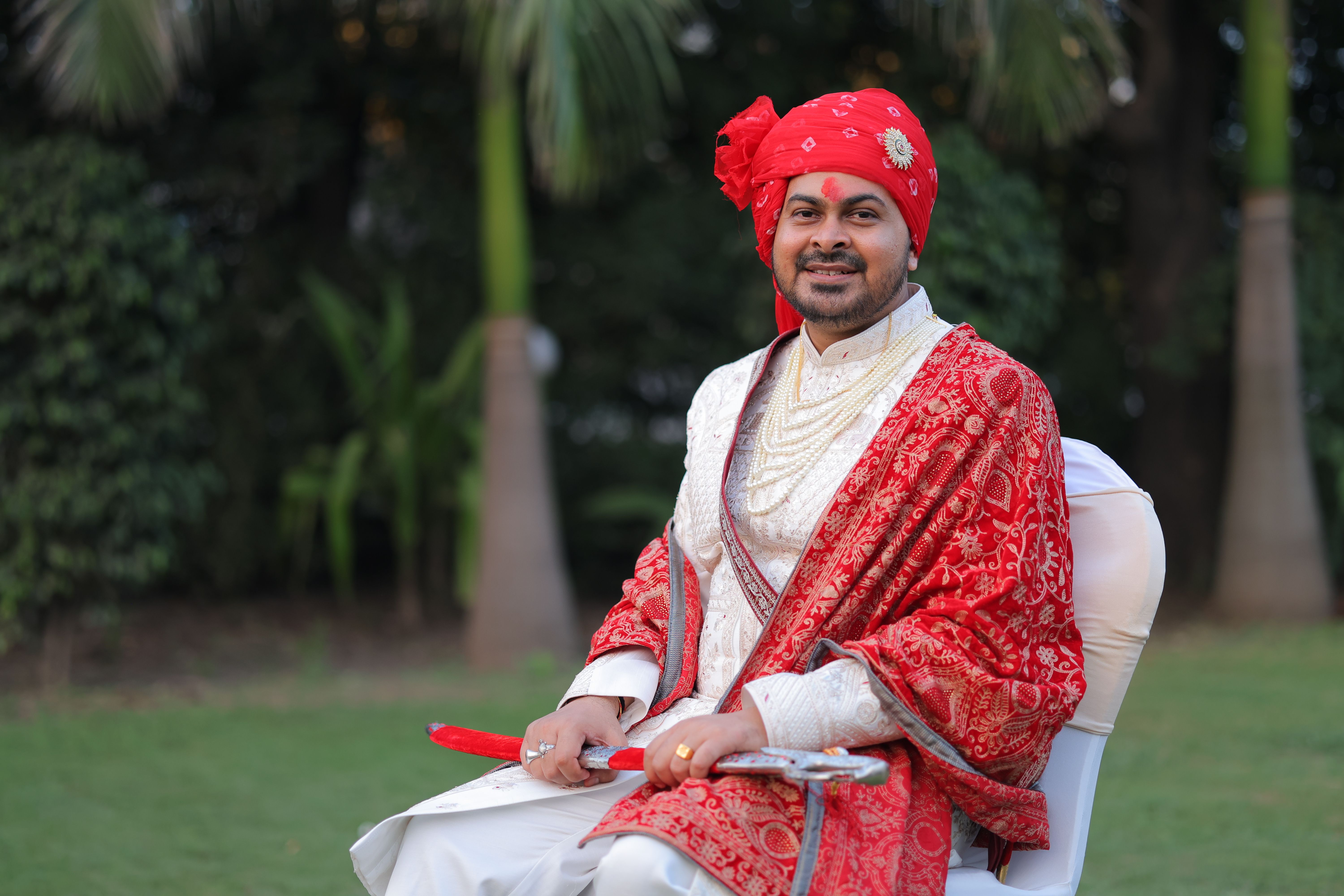 Portrait of a traditional Indian groom sitting outdoors in vibrant red attire, showcasing cultural elegance.