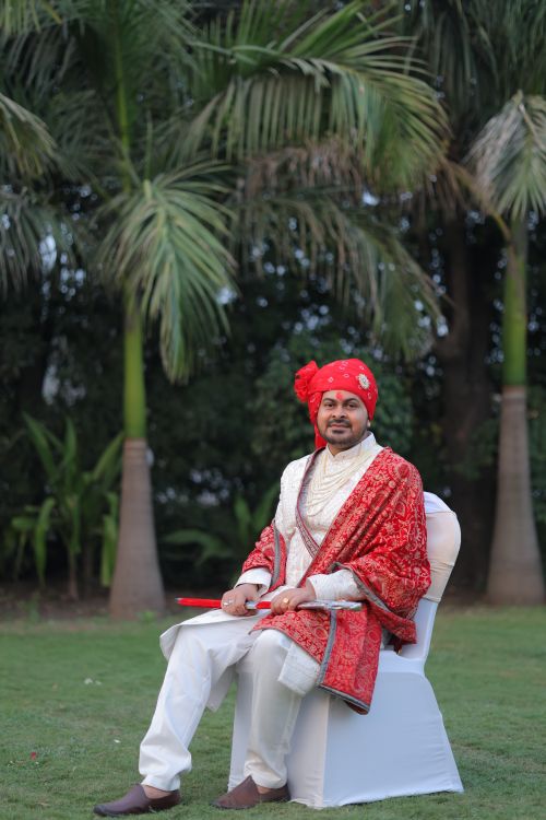 Free Elegant portrait of an Indian groom in traditional red attire seated outdoors with lush greenery. Stock Photo