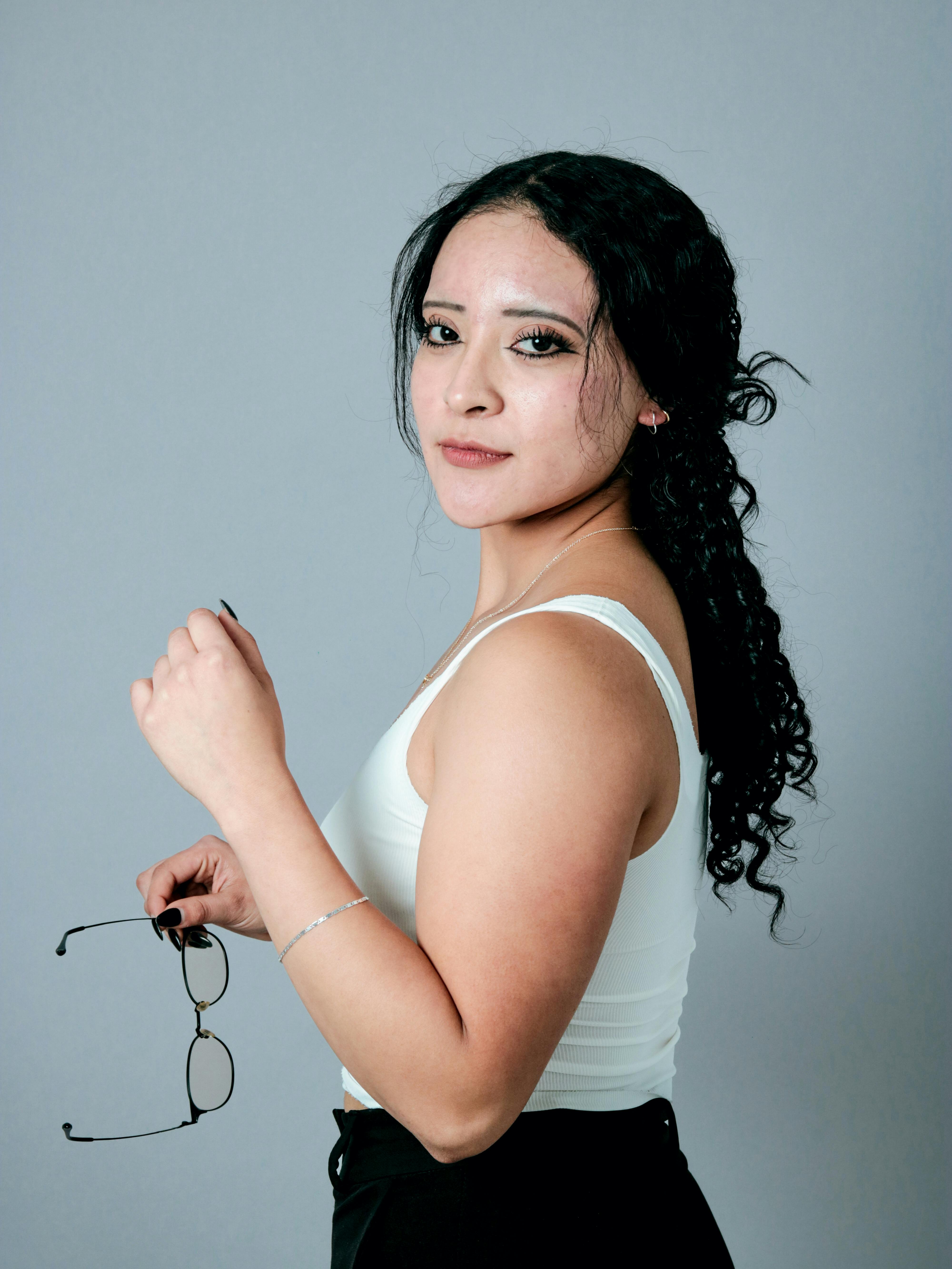 Gratis Ritratto di una donna sicura di sé che tiene in mano degli occhiali e indossa un top bianco su uno sfondo grigio. Foto a disposizione