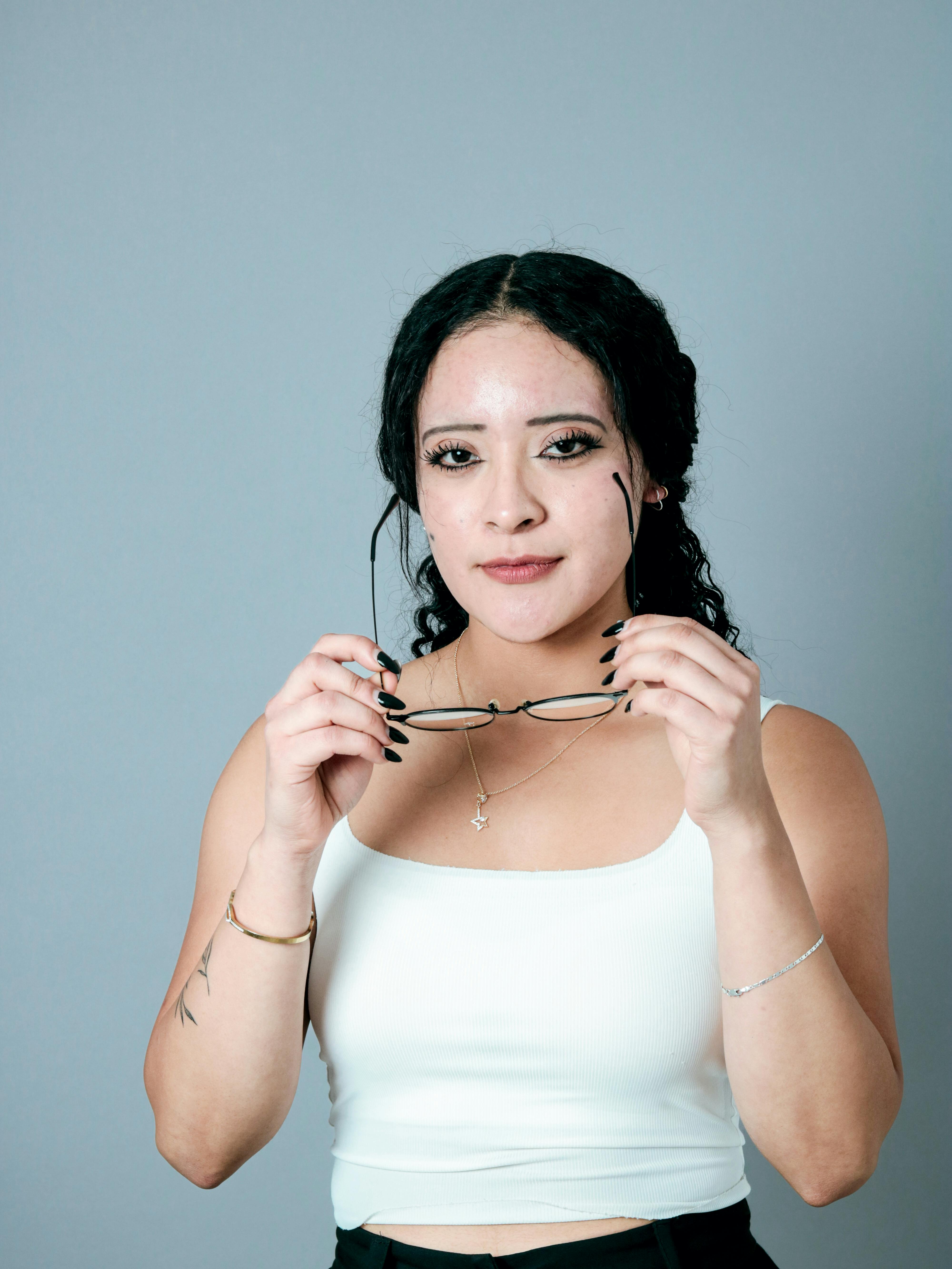 Bezpłatne Młoda kobieta w białym topie pewnie trzyma okulary, wyrażając w ten sposób swoją siłę i styl na neutralnym tle. Zdjęcie z galerii