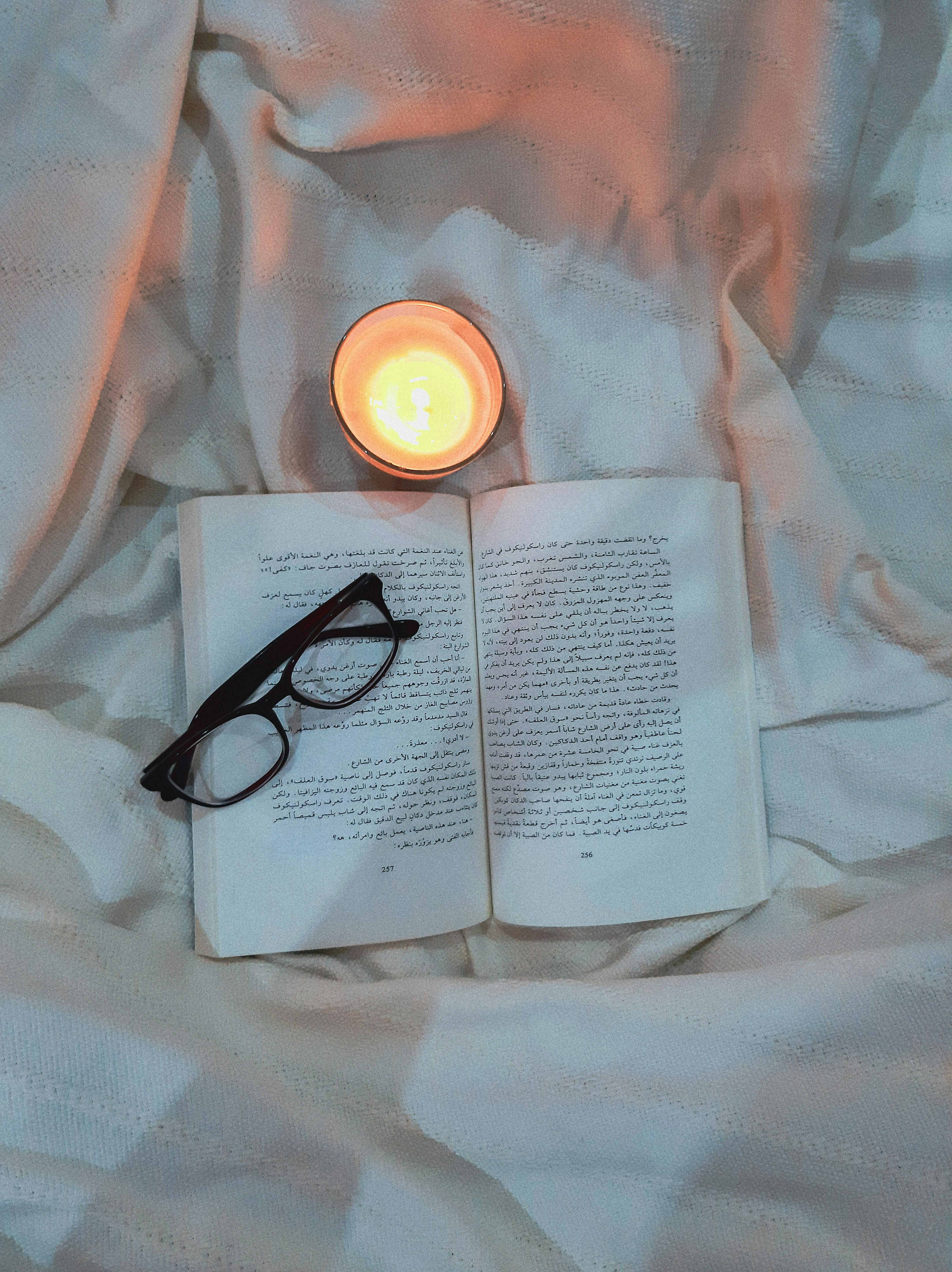 Kostenlos Eine gemütliche Szene mit einem Buch, einer Brille und einer brennenden Kerze auf einer weichen Unterlage. Stock-Foto