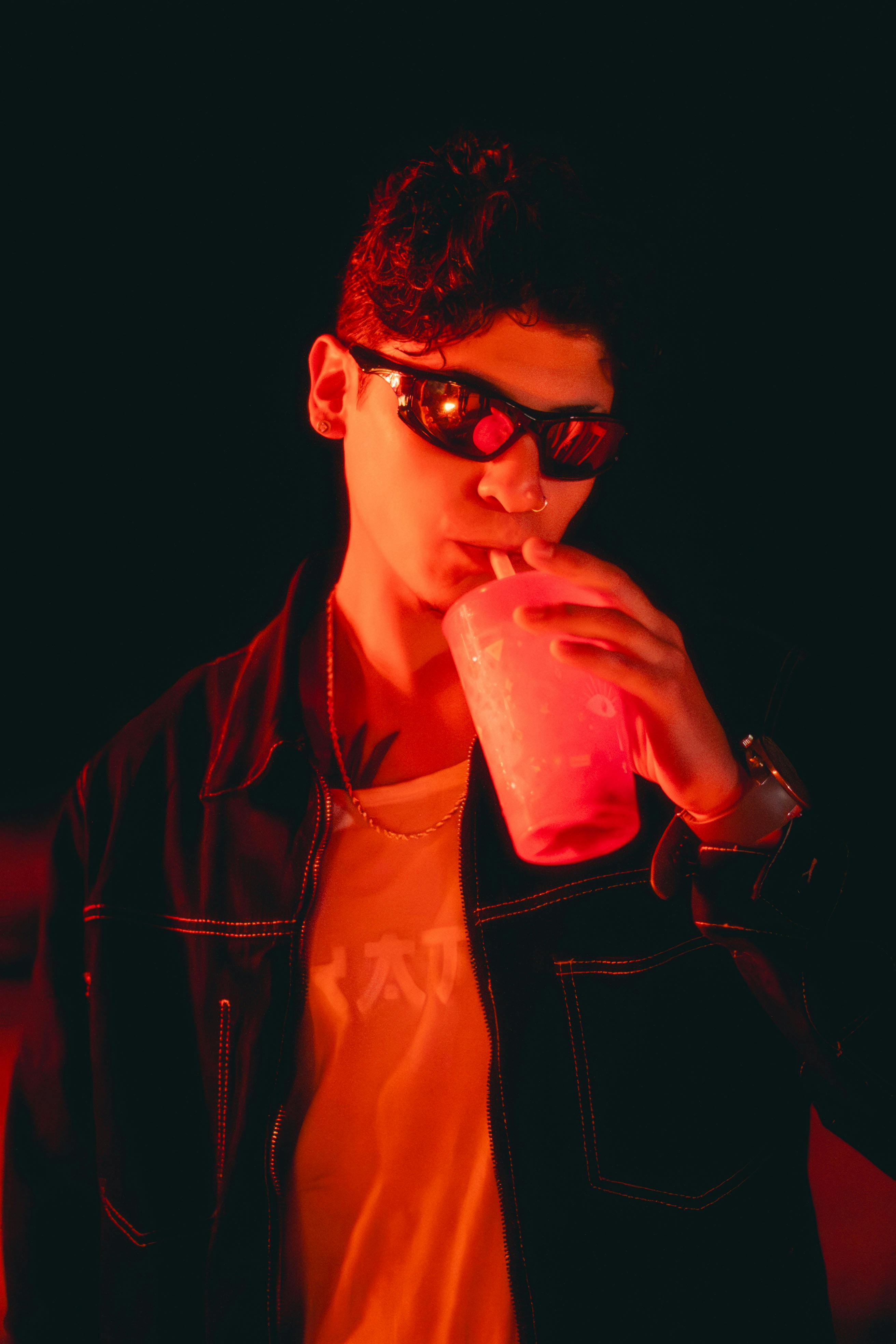 Ücretsiz Güneş gözlüğü takmış, modaya uygun giyinmiş bir genç kız, canlı neon ışıklar altında içeceğinin tadını çıkarıyor. Stok Fotoğraflar