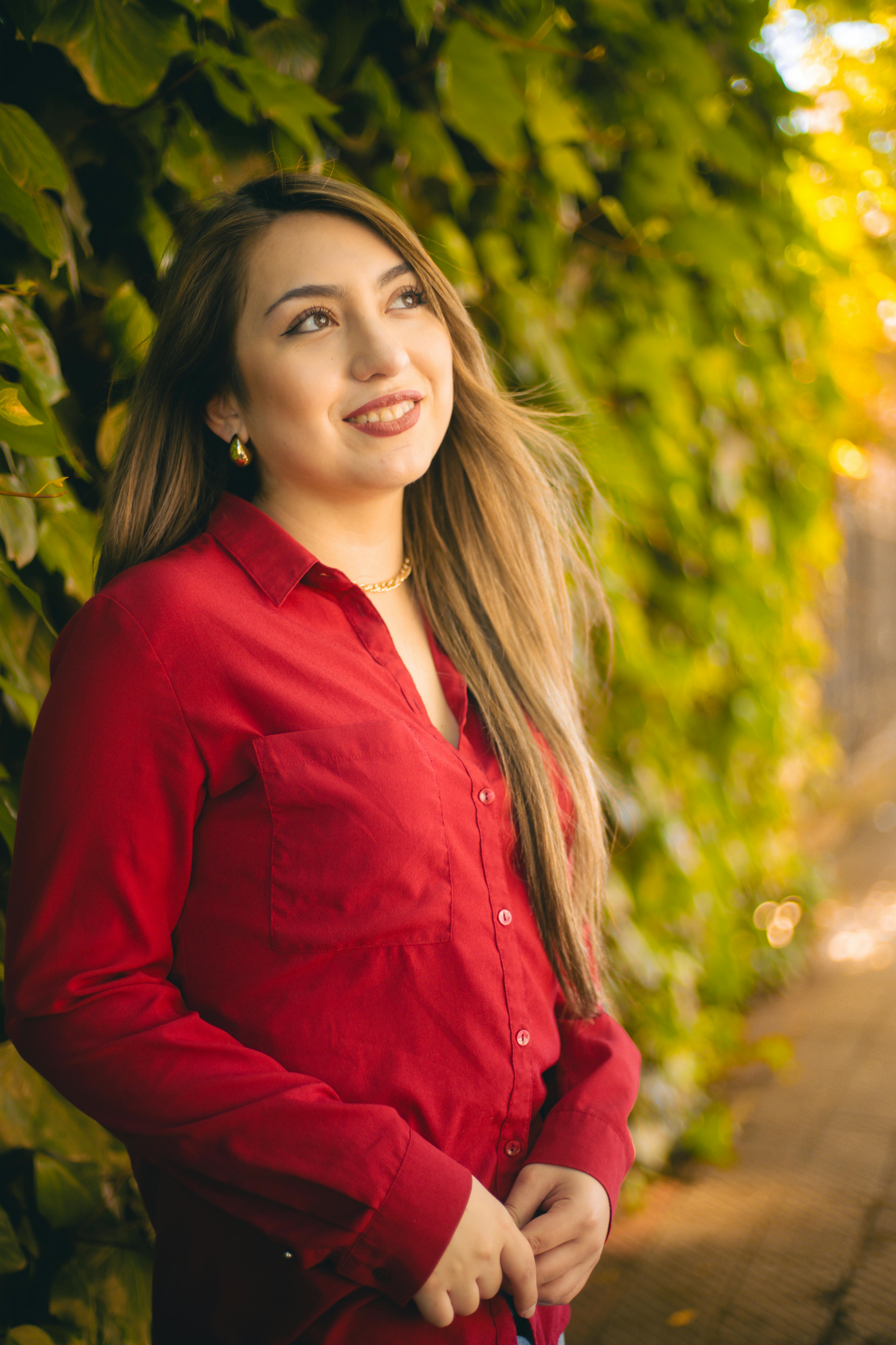Free Smiling woman in red shirt against vibrant green leaves, enjoying a sunny day. Stock Photo