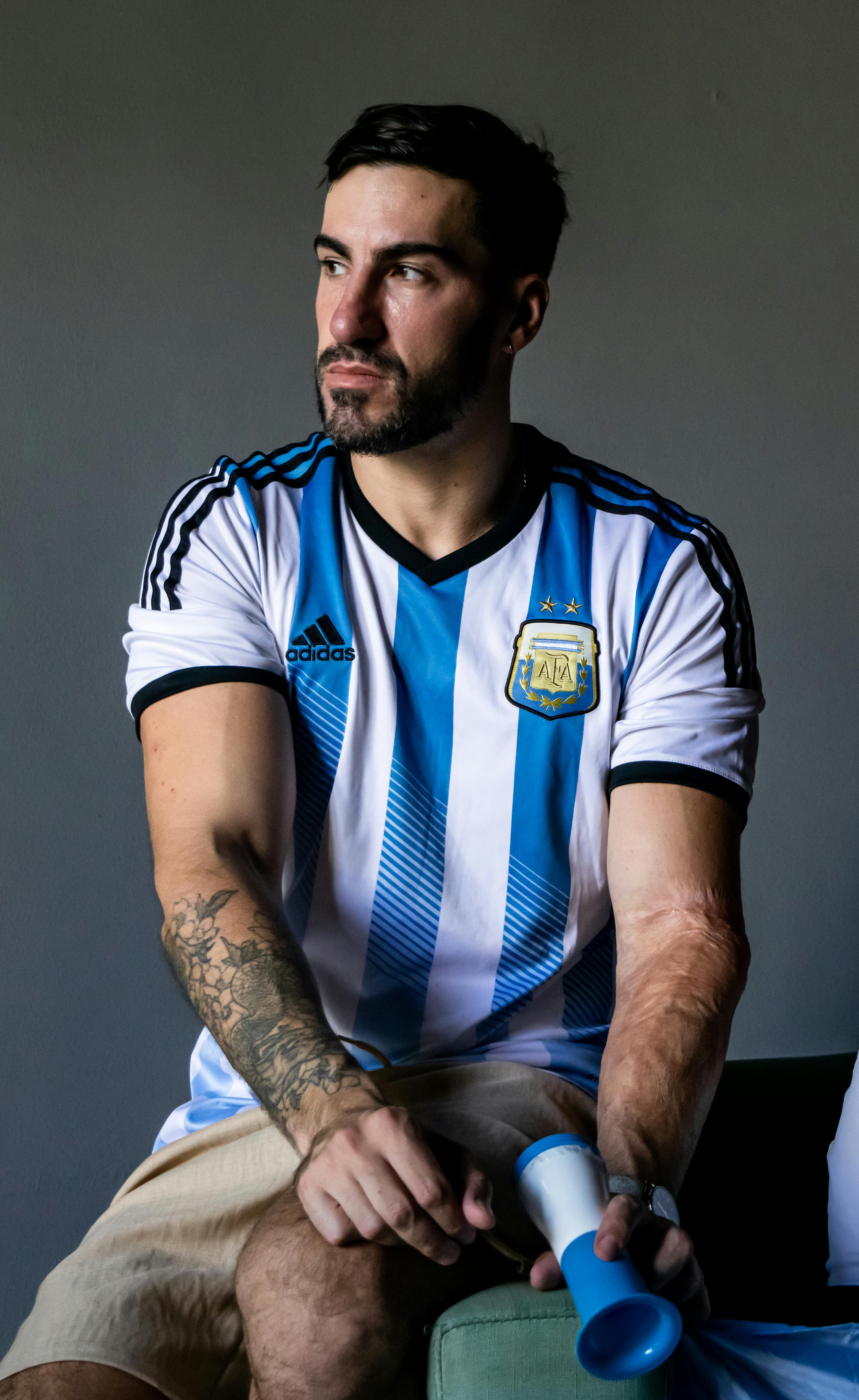Adult man sits wearing an Argentina football jersey holding a blue megaphone, gaze turned sideways.