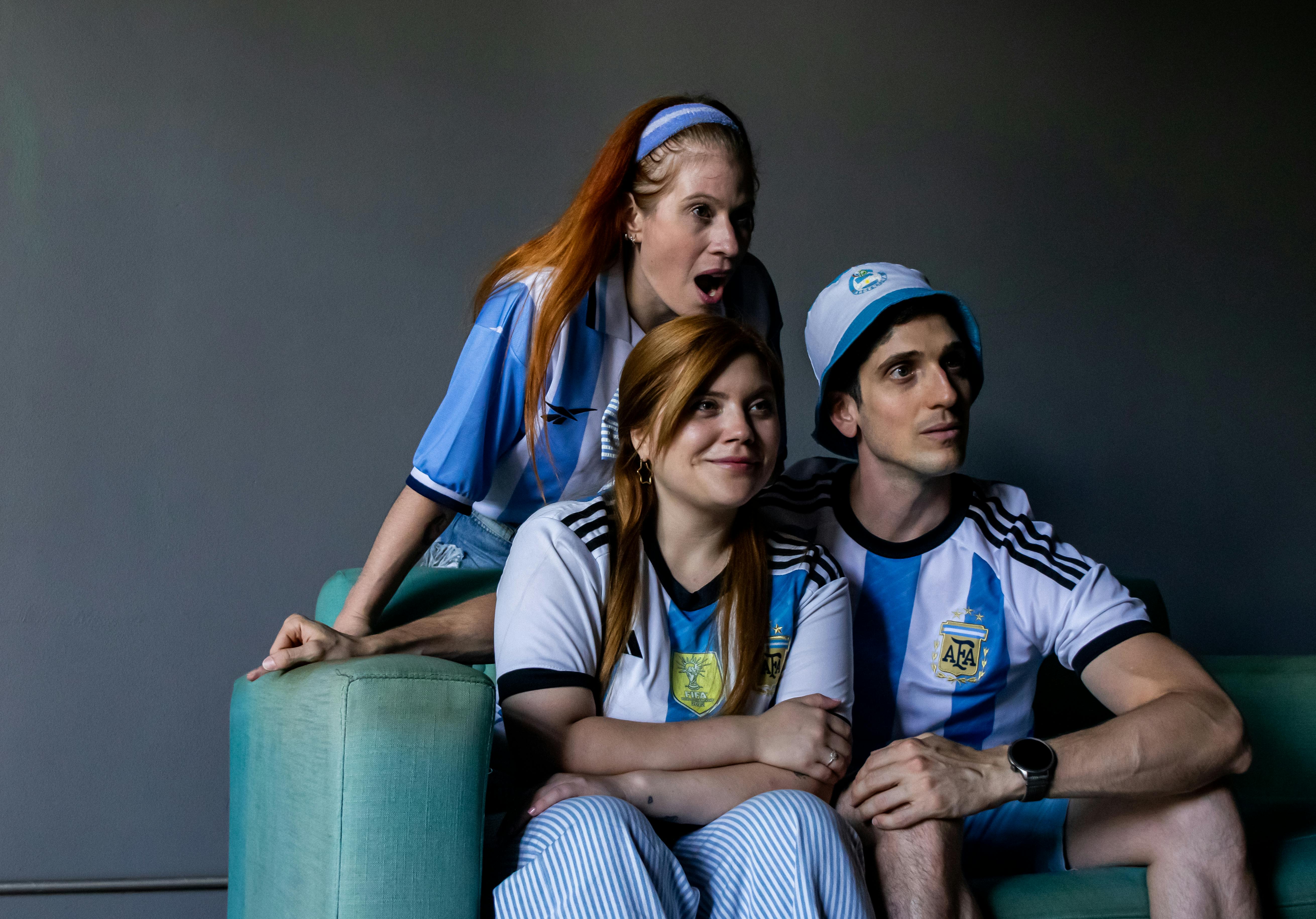 Three fans in Argentina jerseys watch a soccer match with excitement indoors.
