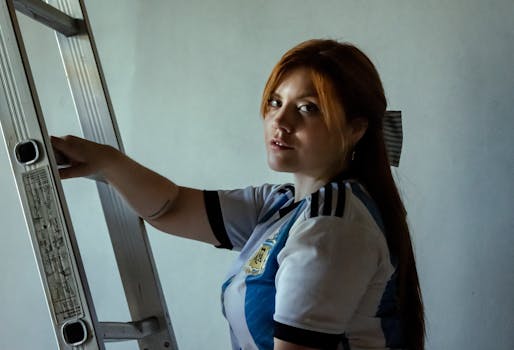 Young woman in an Argentina football jersey standing beside a ladder indoors, looking thoughtful.