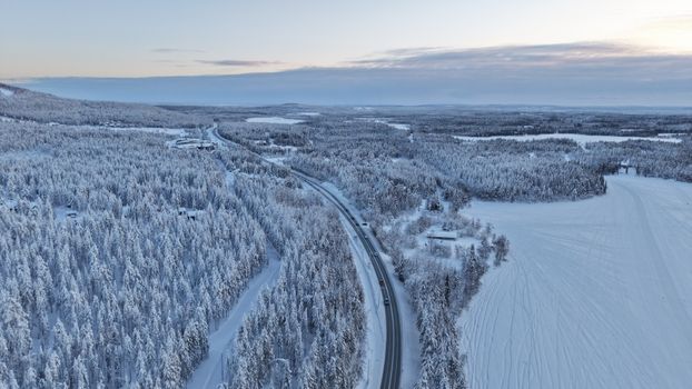 Aerial shot of a snowy road winding through a winter forest in Finland, at sunset.