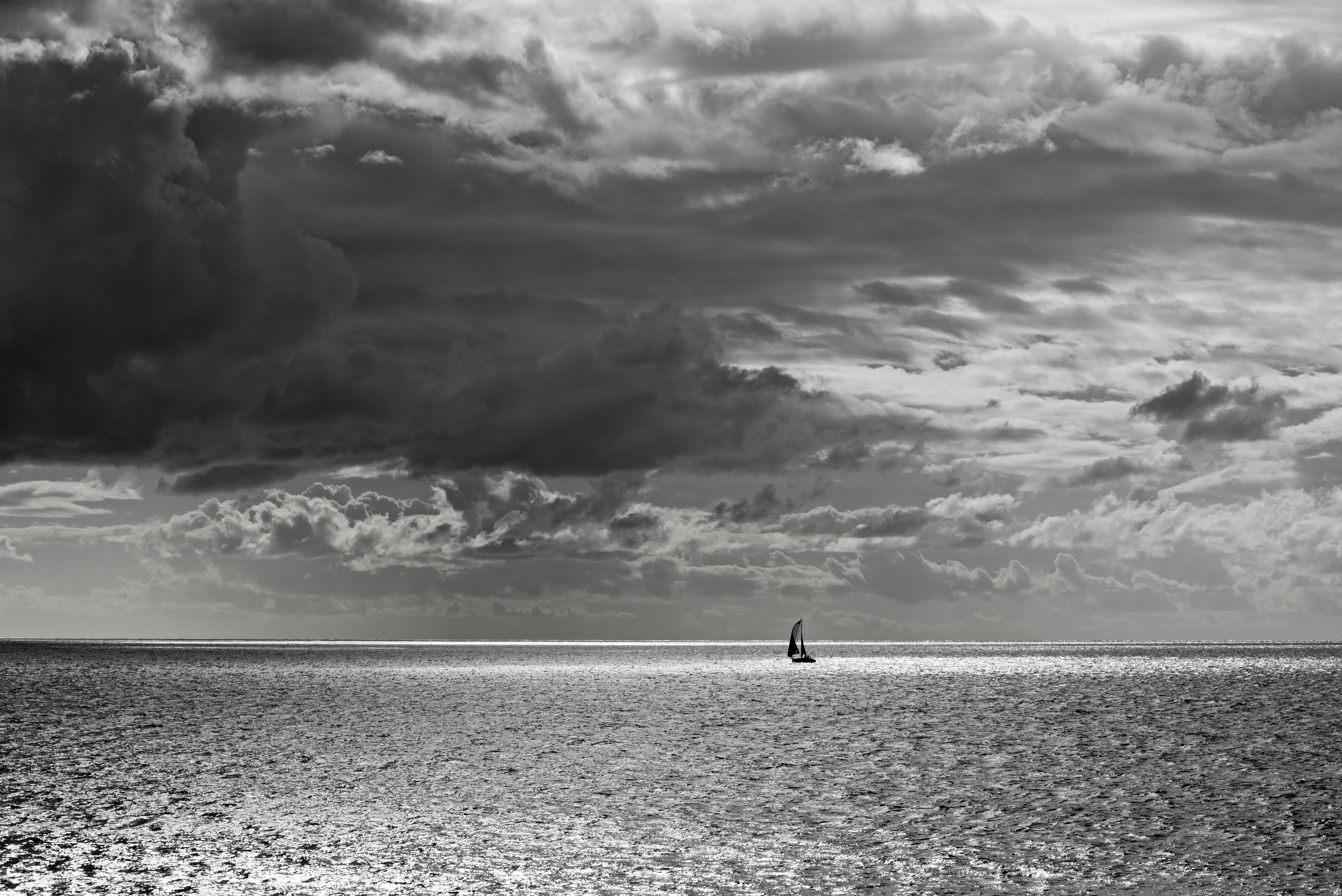 Free A lone sailboat navigates a vast ocean under dramatic skies in stunning black and white. Stock Photo