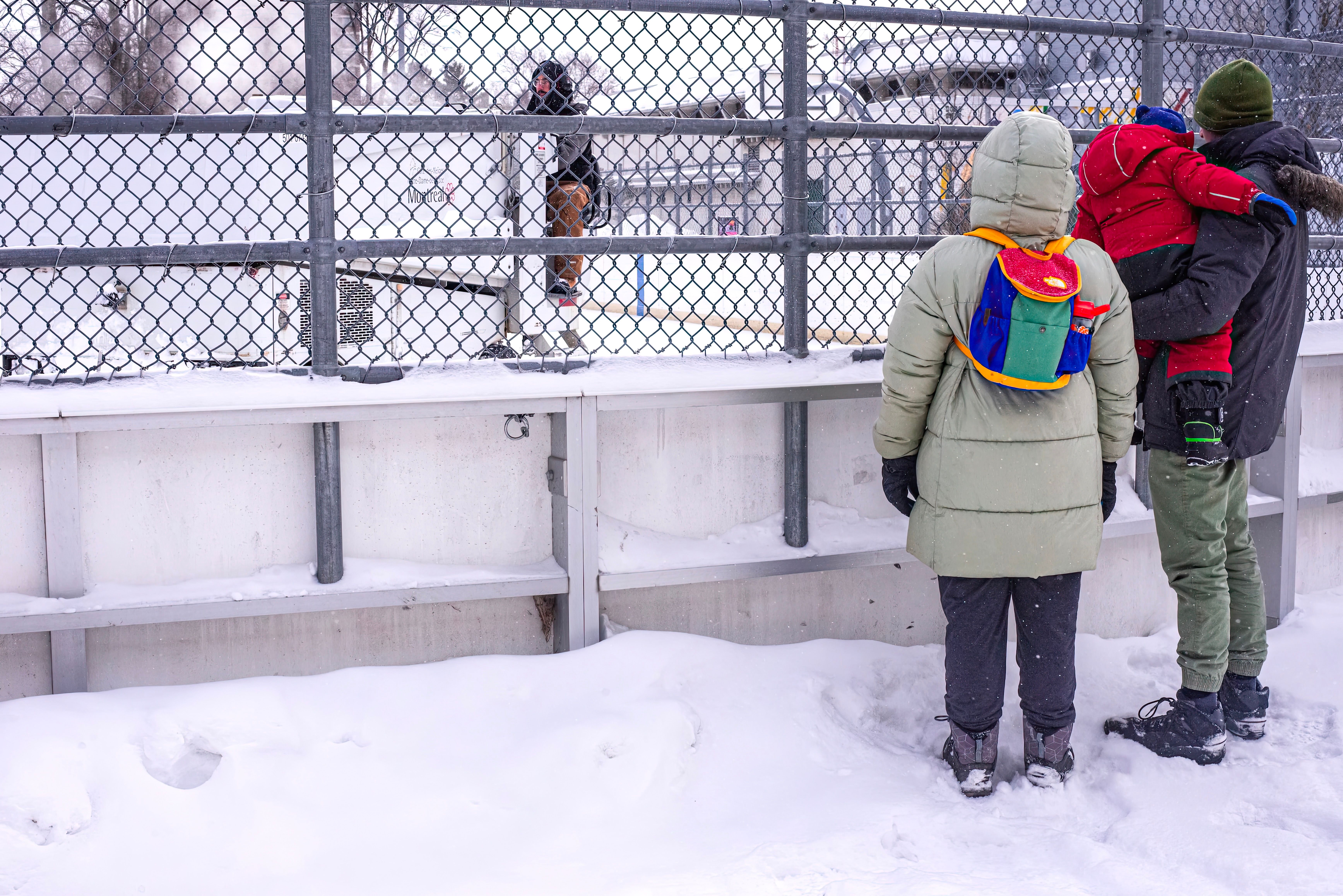 A family bundled in winter clothes watches an outdoor hockey game through a fence on a snowy day.