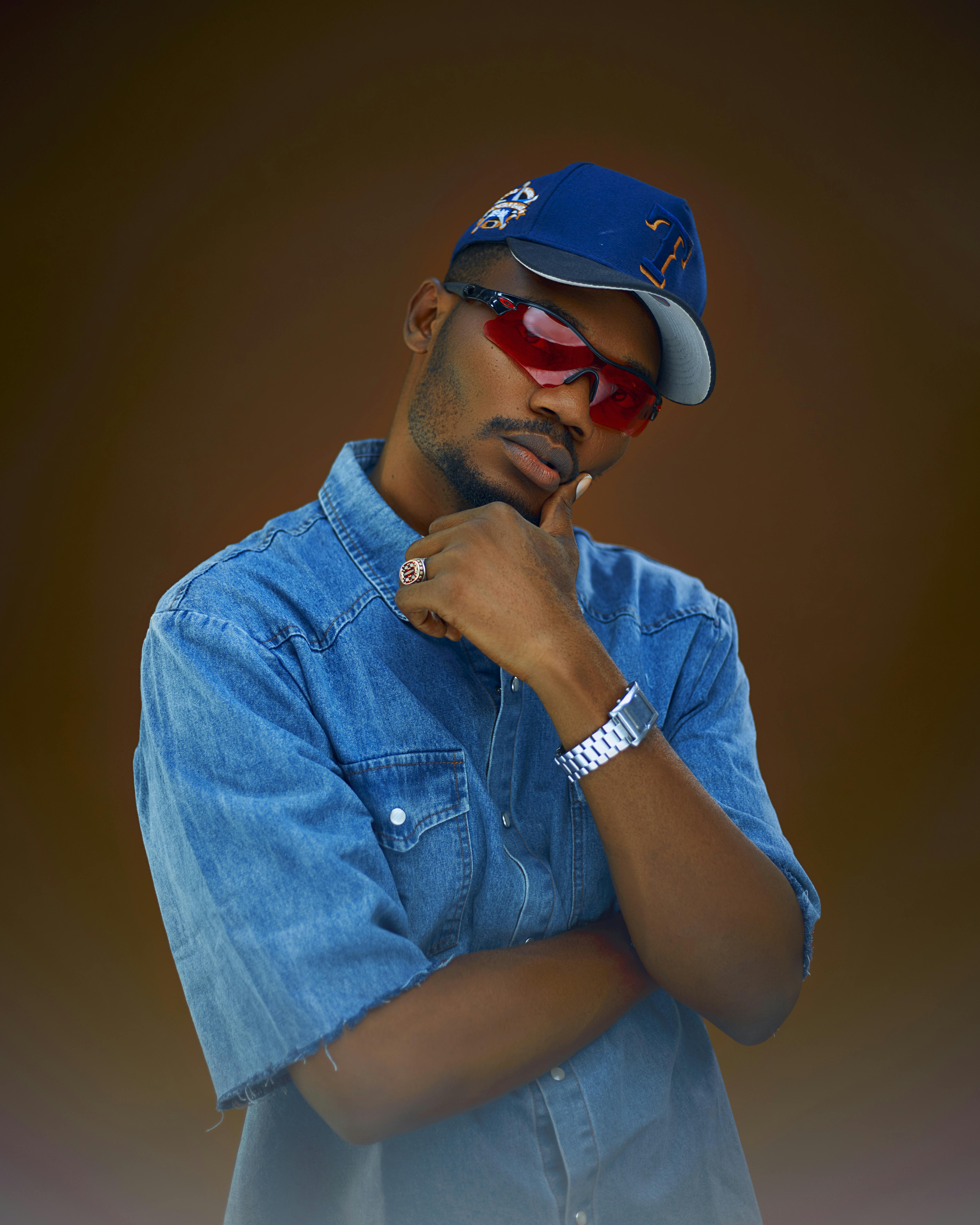 ฟรี ภาพถ่ายบุคคลของชายคนหนึ่งในชุดยีนส์ สวมหมวกและแว่นกันแดด โพสท่าอย่างมั่นใจ คลังภาพถ่าย