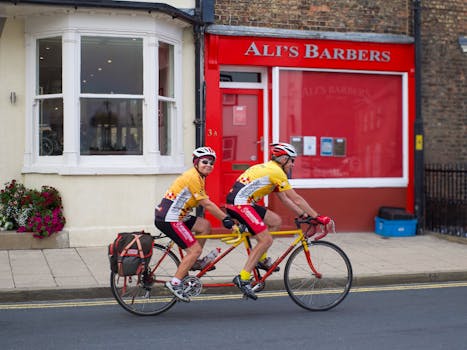 Two cyclists ride a tandem bike past Ali's Barbers in Boroughbridge, UK.