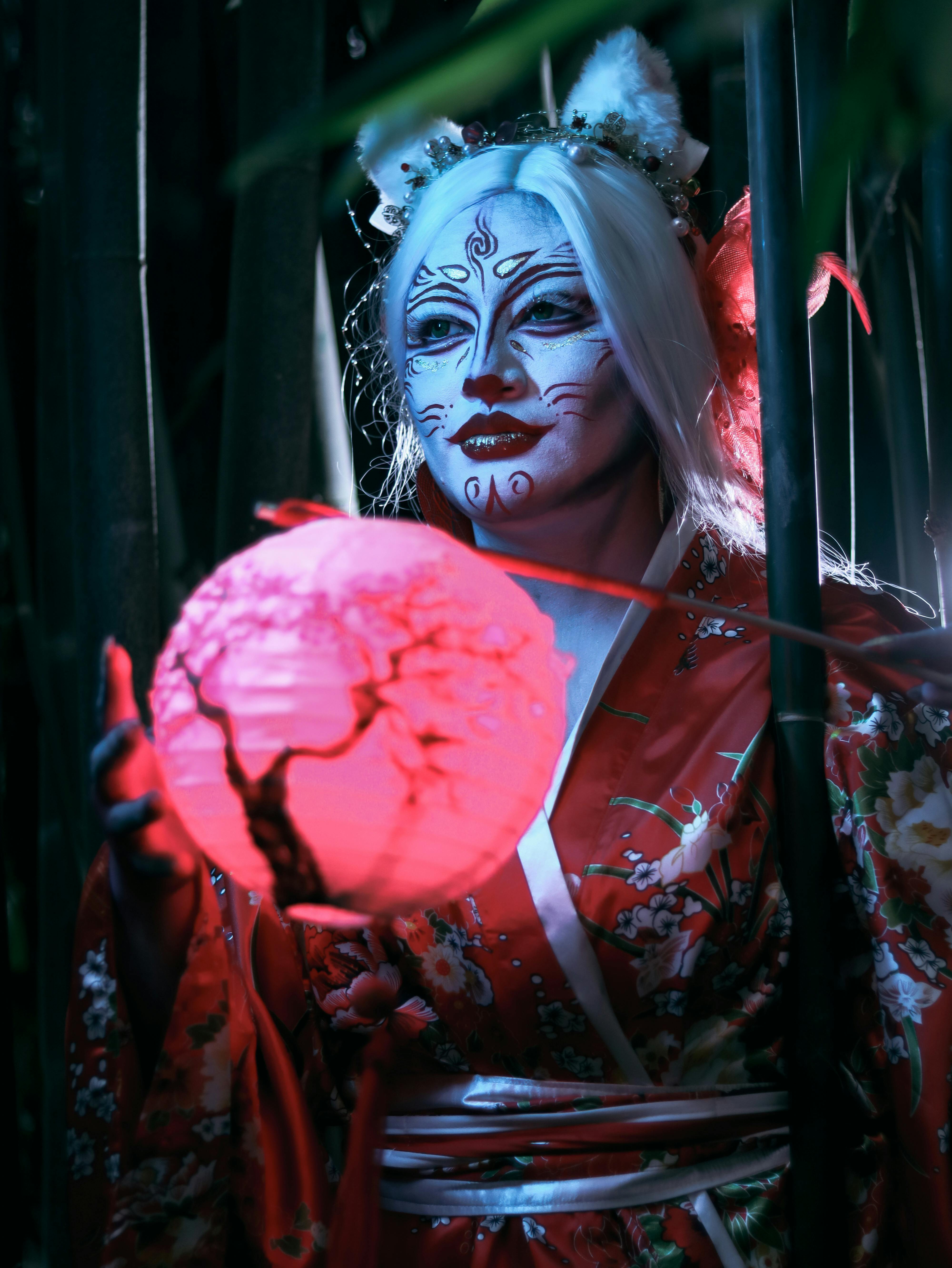 De franc Una dona amb un quimono floral i pintura facial sosté una llanterna brillant en un bosquet de bambús. Foto d'estoc