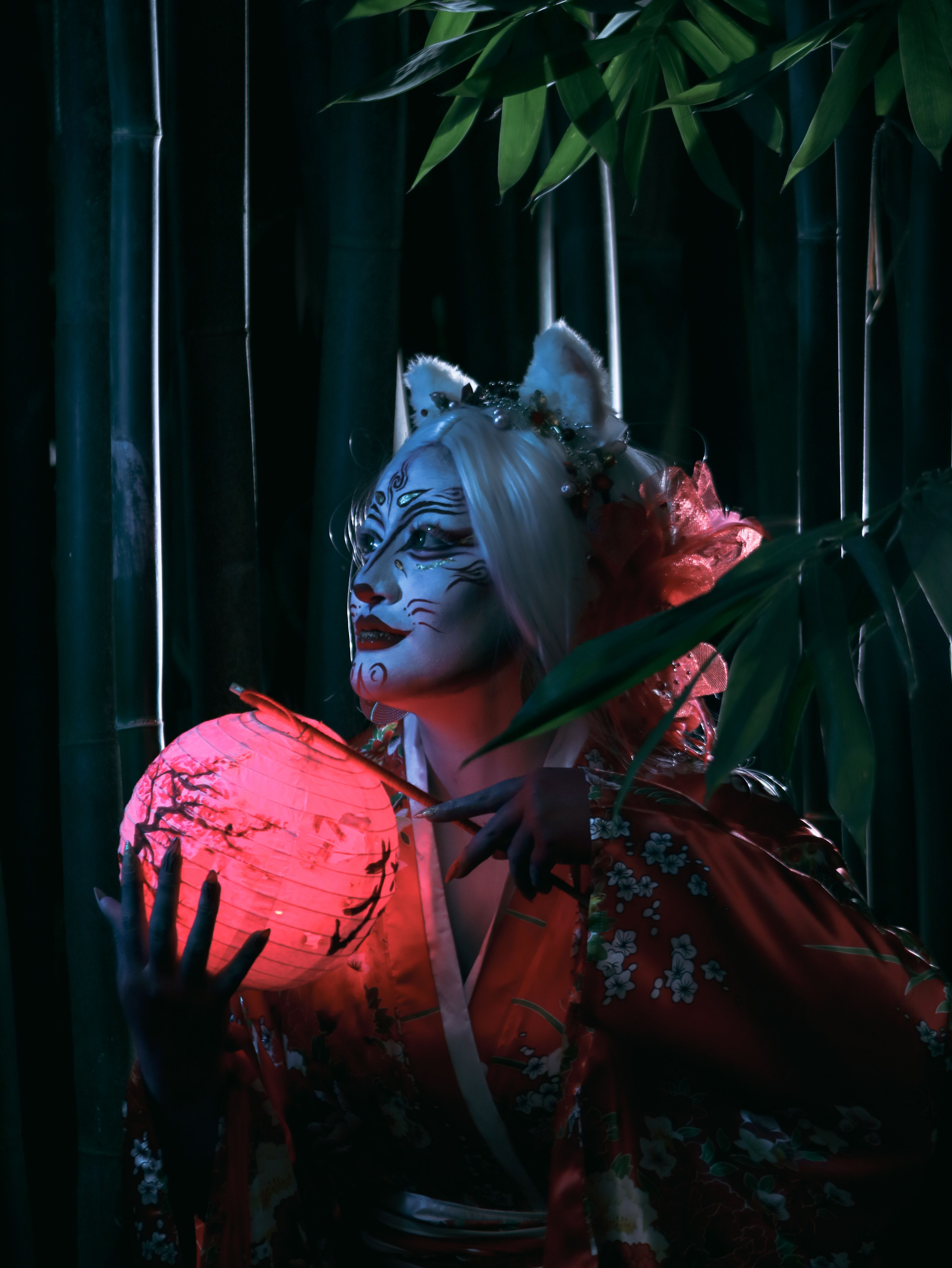 A performer in kimono with face paint holding a red lantern in a bamboo forest