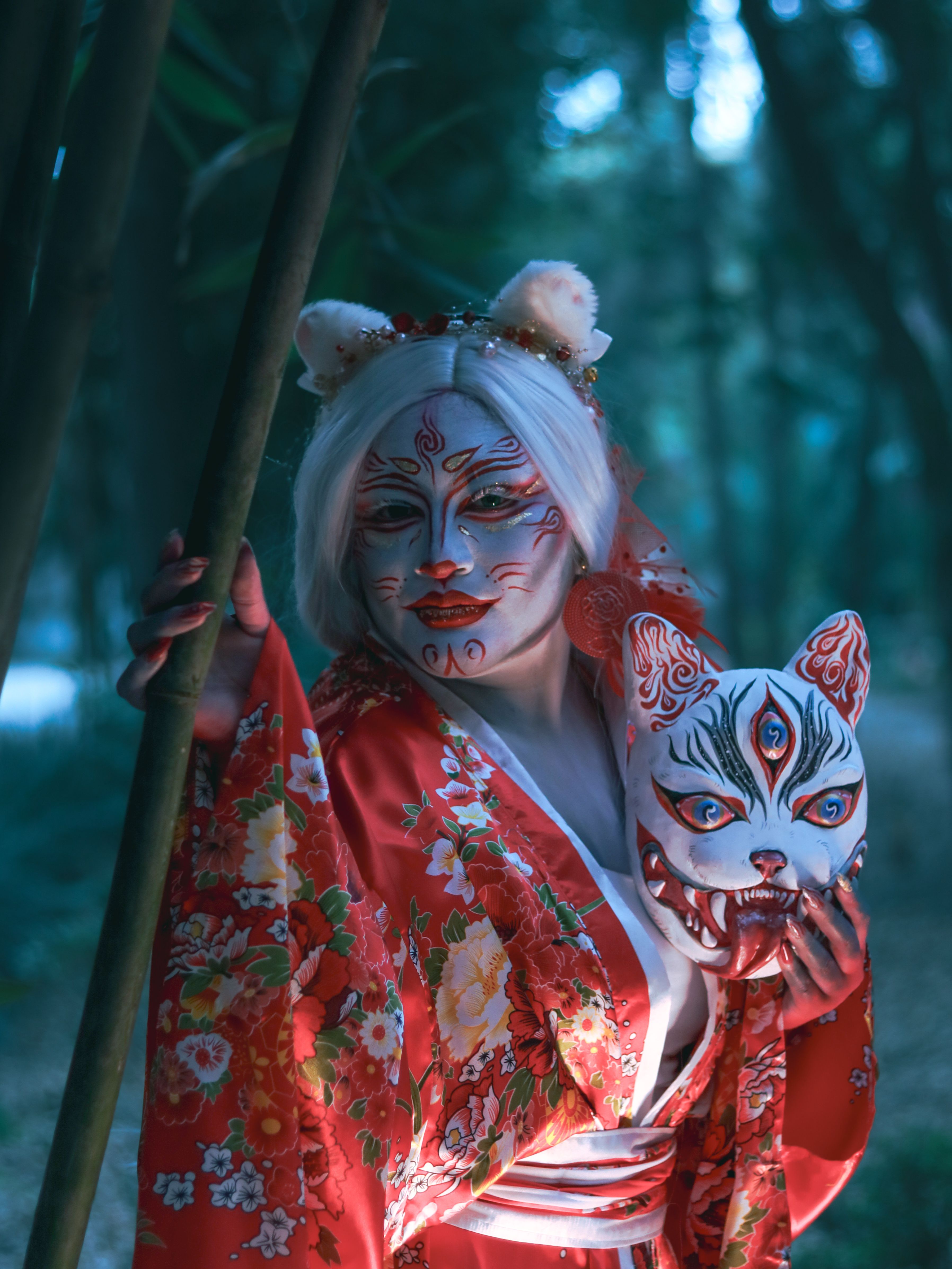 A captivating cosplay with kitsune mask, floral kimono, and intricate face paint in a forested setting.