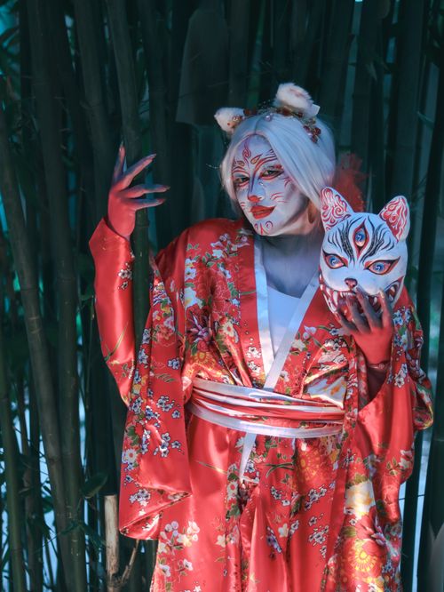 Free A person in an ornate red kimono holding a mystical fox mask in a bamboo setting. Stock Photo