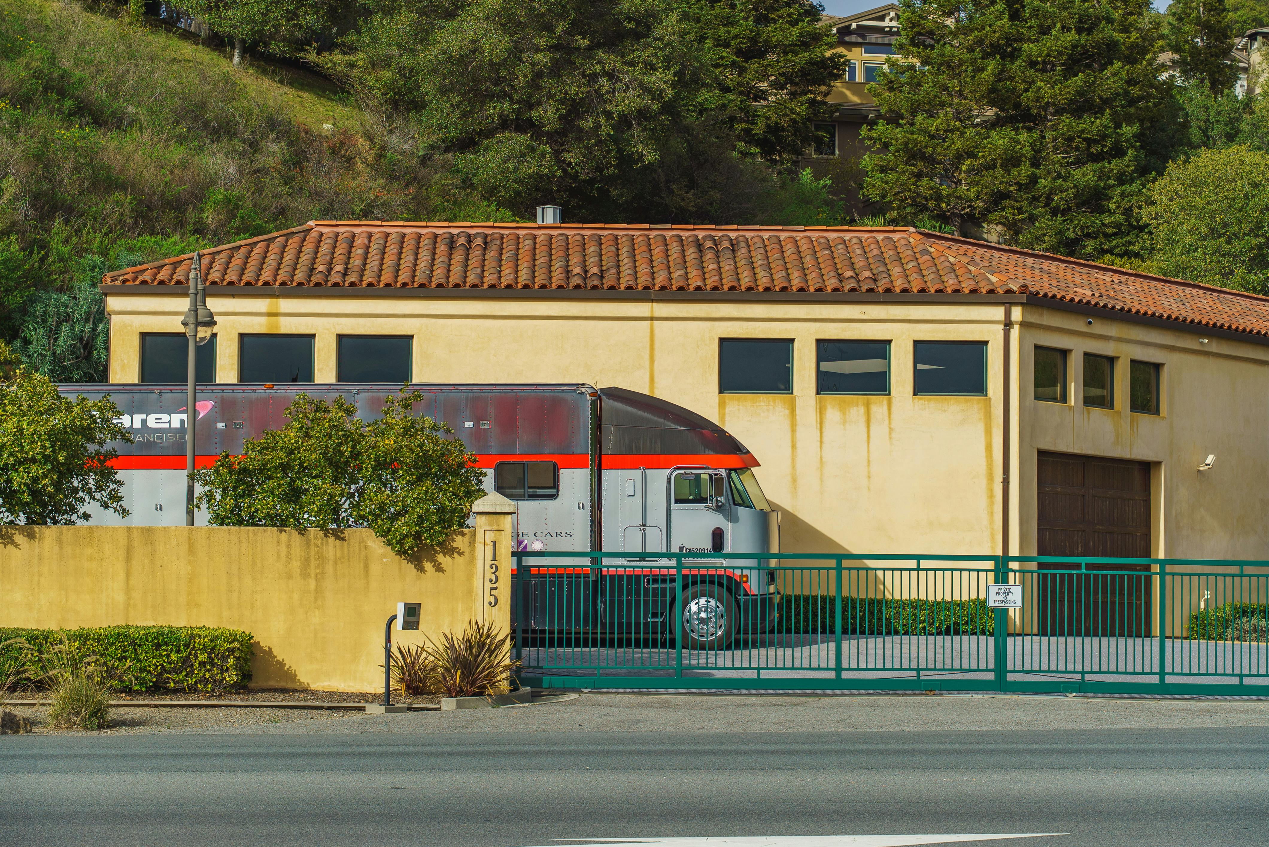 Miễn phí Một chiếc xe tải với các điểm nhấn màu đỏ đang đậu bên ngoài một tòa nhà màu nâu nhạt có mái ngói đỏ, xung quanh là cây xanh. Ảnh lưu trữ