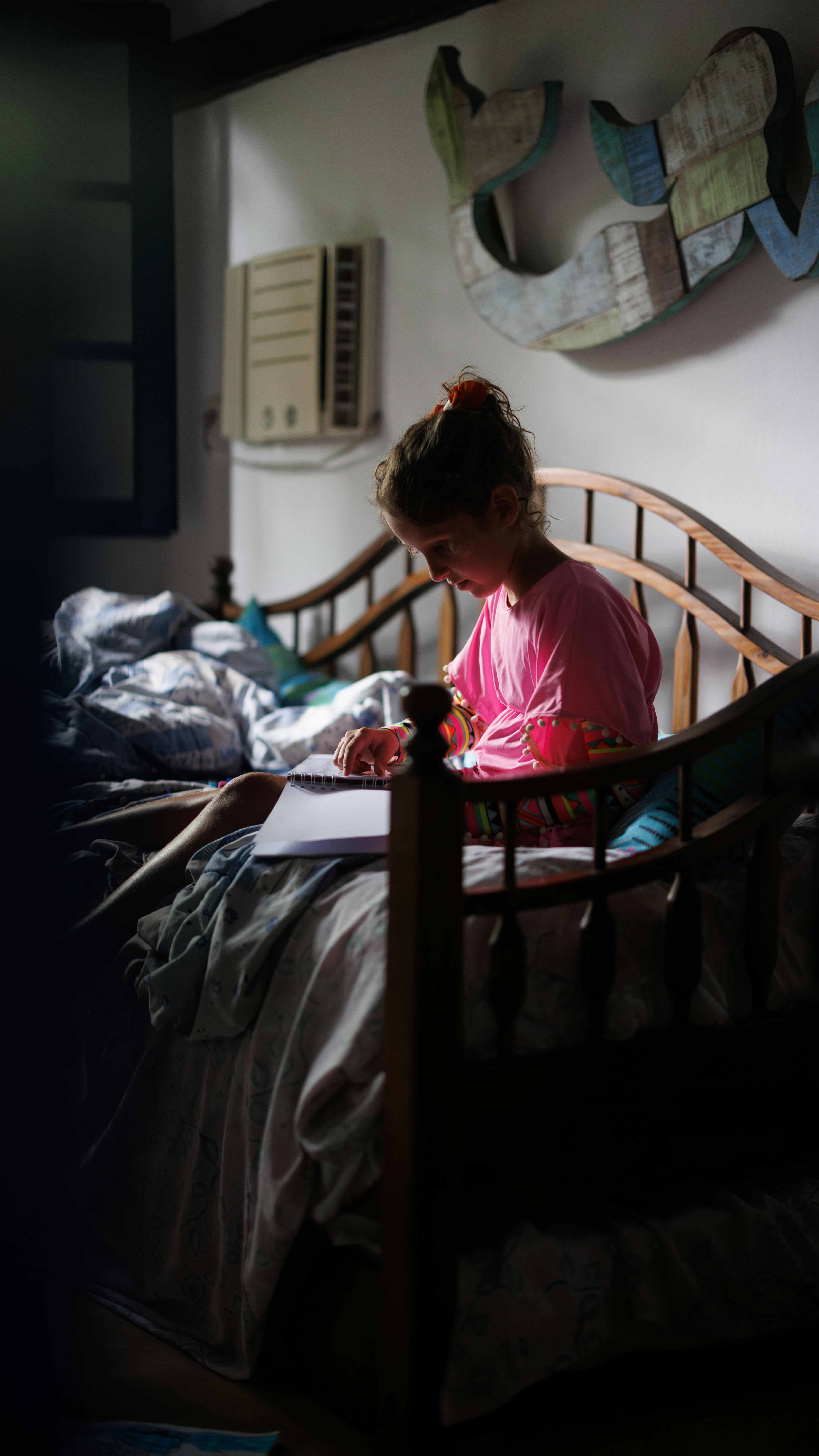 gratis Een jong meisje leest een boek bij het raam in een zacht verlichte slaapkamer, wat een contemplatieve sfeer creëert. Stockfoto