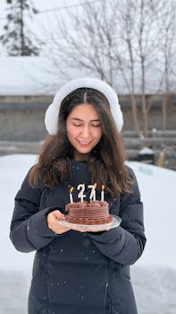 Smiling woman holding a birthday cake with candles in a snowy outdoor setting.