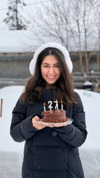 Young woman holding birthday cake with candles, smiling outdoors in winter snow scene.
