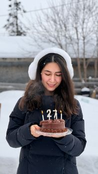 Woman celebrating her birthday outdoors in winter with a chocolate cake and candles.