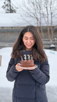 Smiling woman holding a chocolate birthday cake with candles in a snowy outdoor setting.