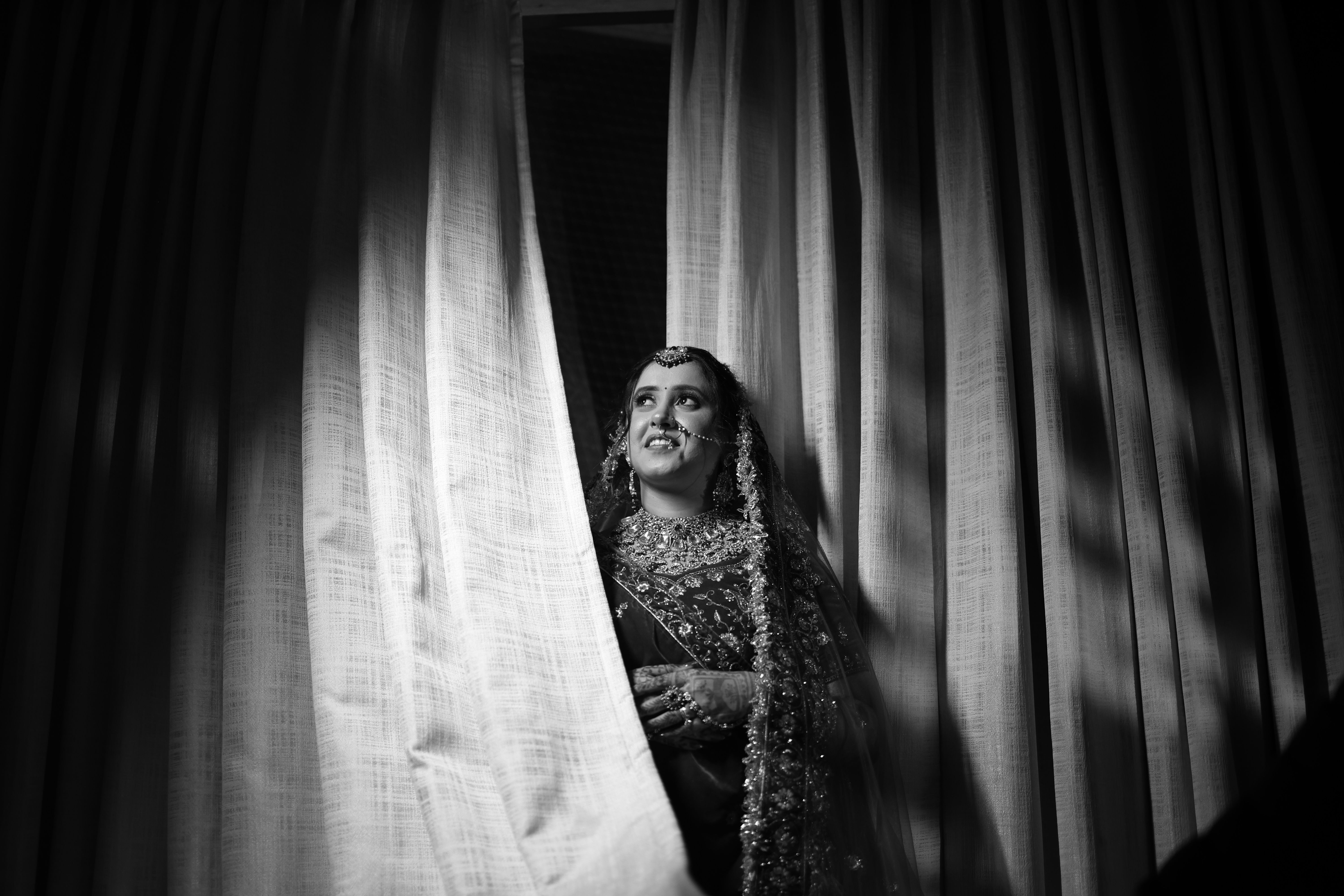 Beautiful black and white portrait of an Indian bride in traditional attire by the curtain.