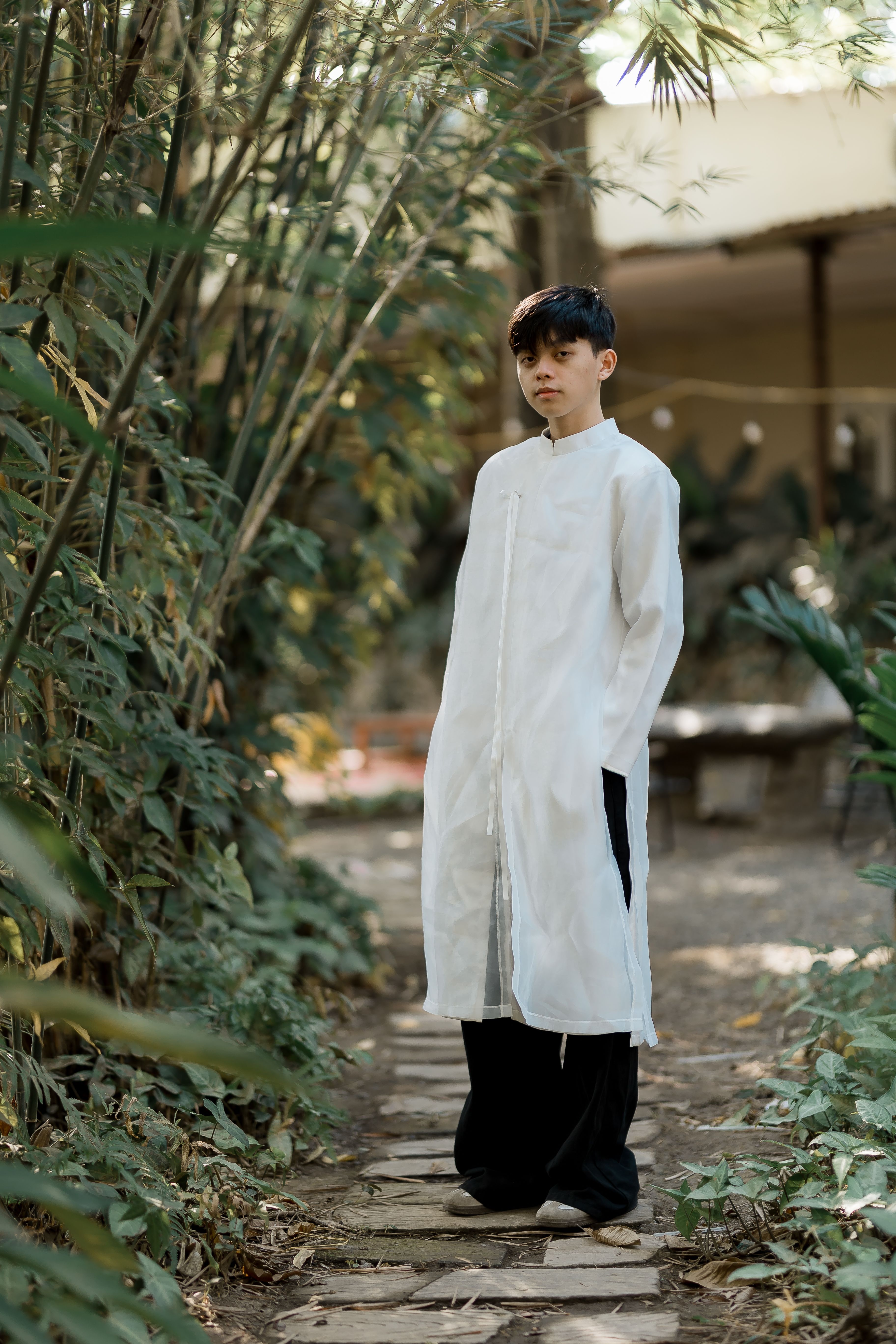 A young man in traditional white attire standing on a garden path with lush greenery around.
