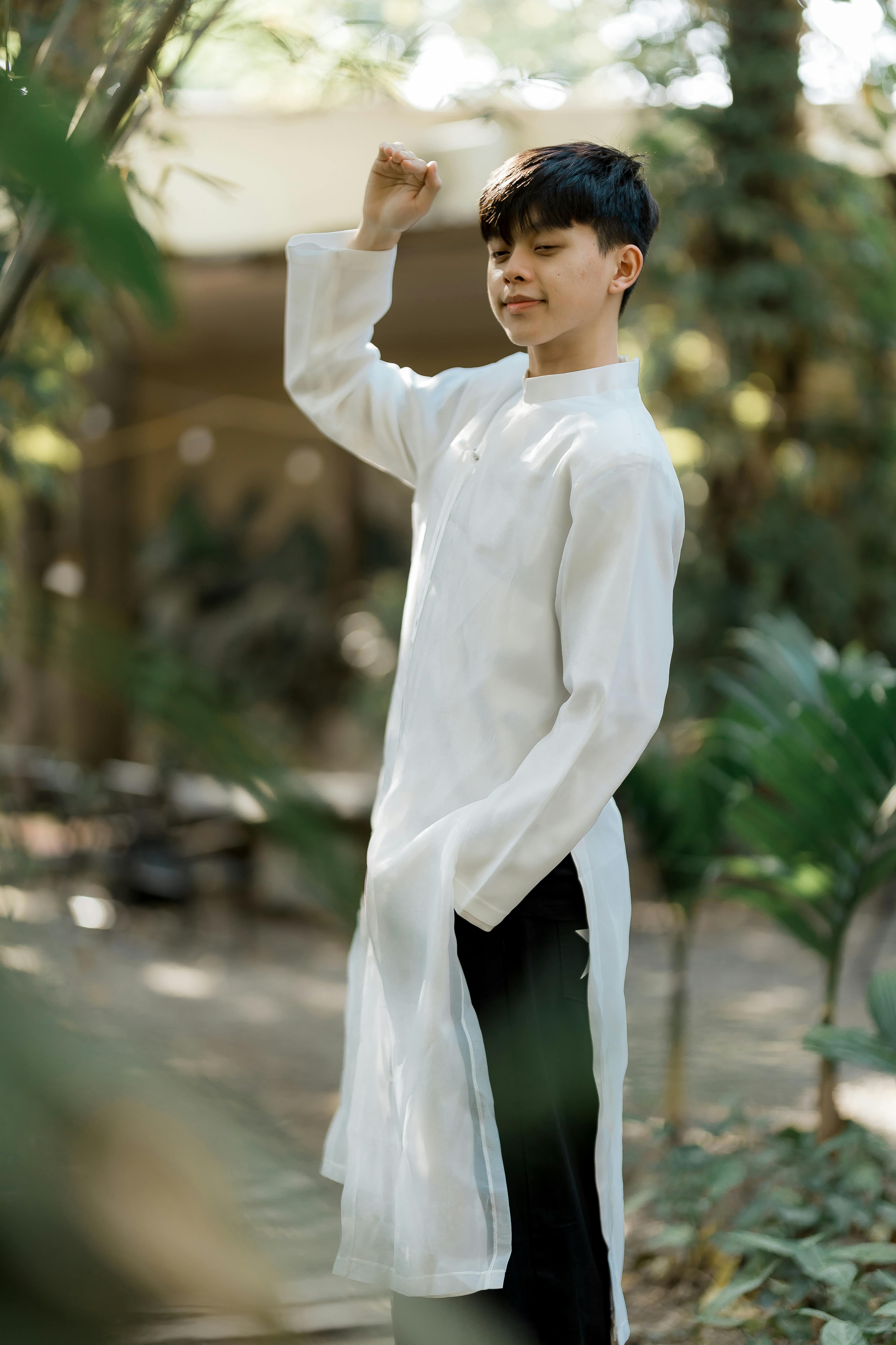 Gratis Un giovane uomo in abiti tradizionali bianchi posa con sicurezza in un ambiente esterno circondato dal verde. Foto a disposizione