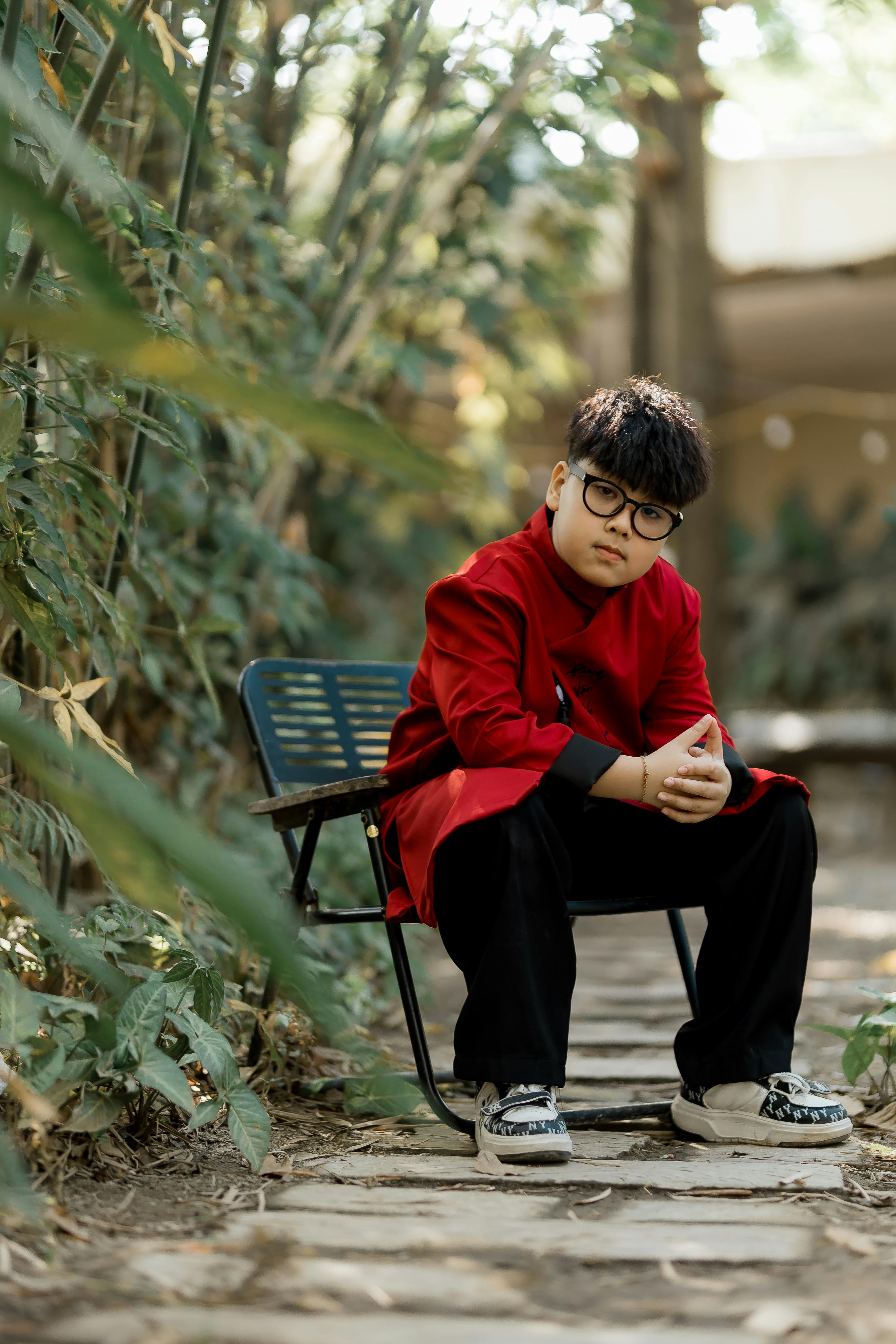 Gratis Un ragazzino che indossa un cappotto rosso è seduto su una sedia, circondato dal verde e dalla luce naturale. Foto a disposizione
