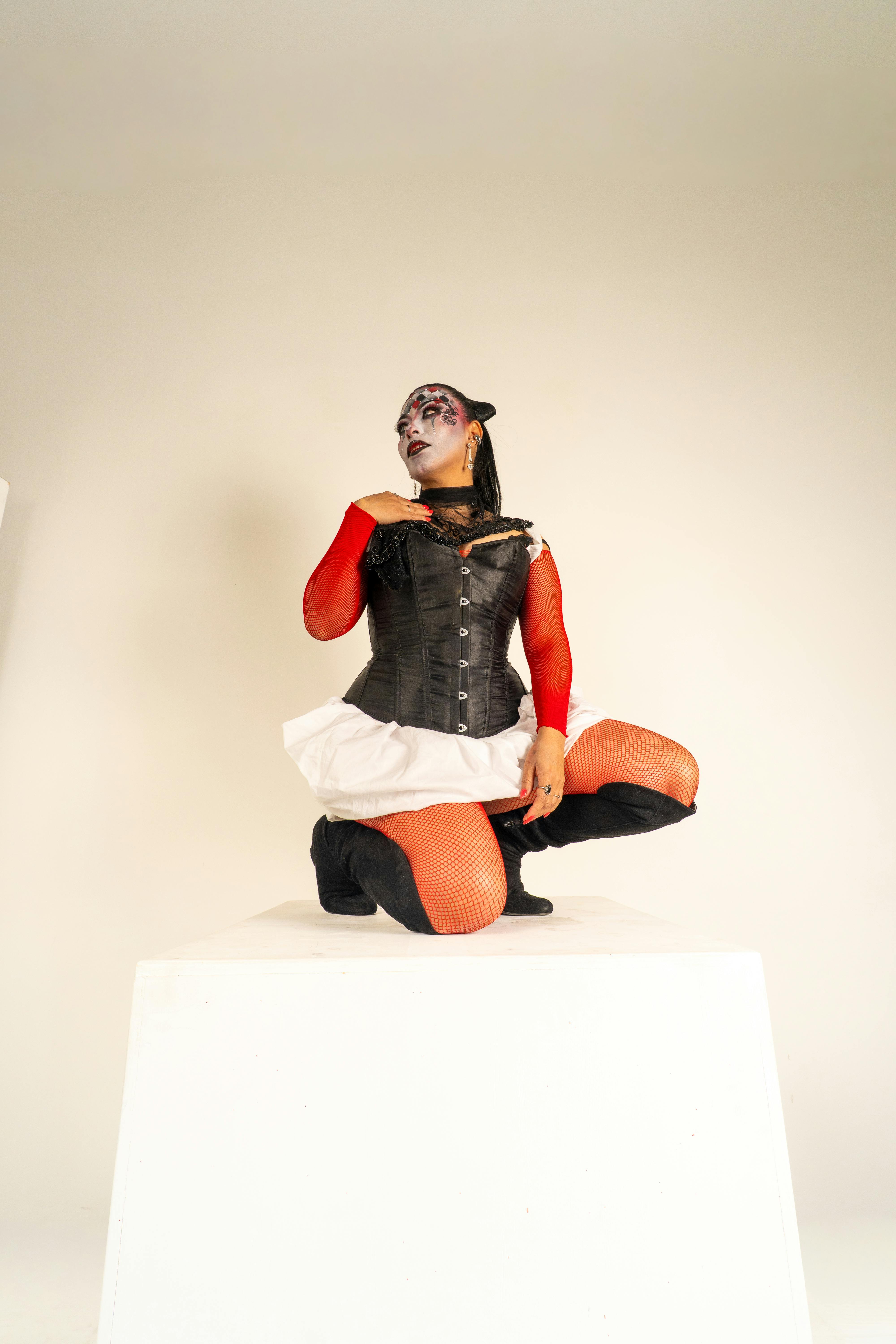 gratis Een Latijns-Amerikaanse vrouw met artistieke clownsschmink knielt op een voetstuk. Opvallende rood-zwarte outfit. Stockfoto