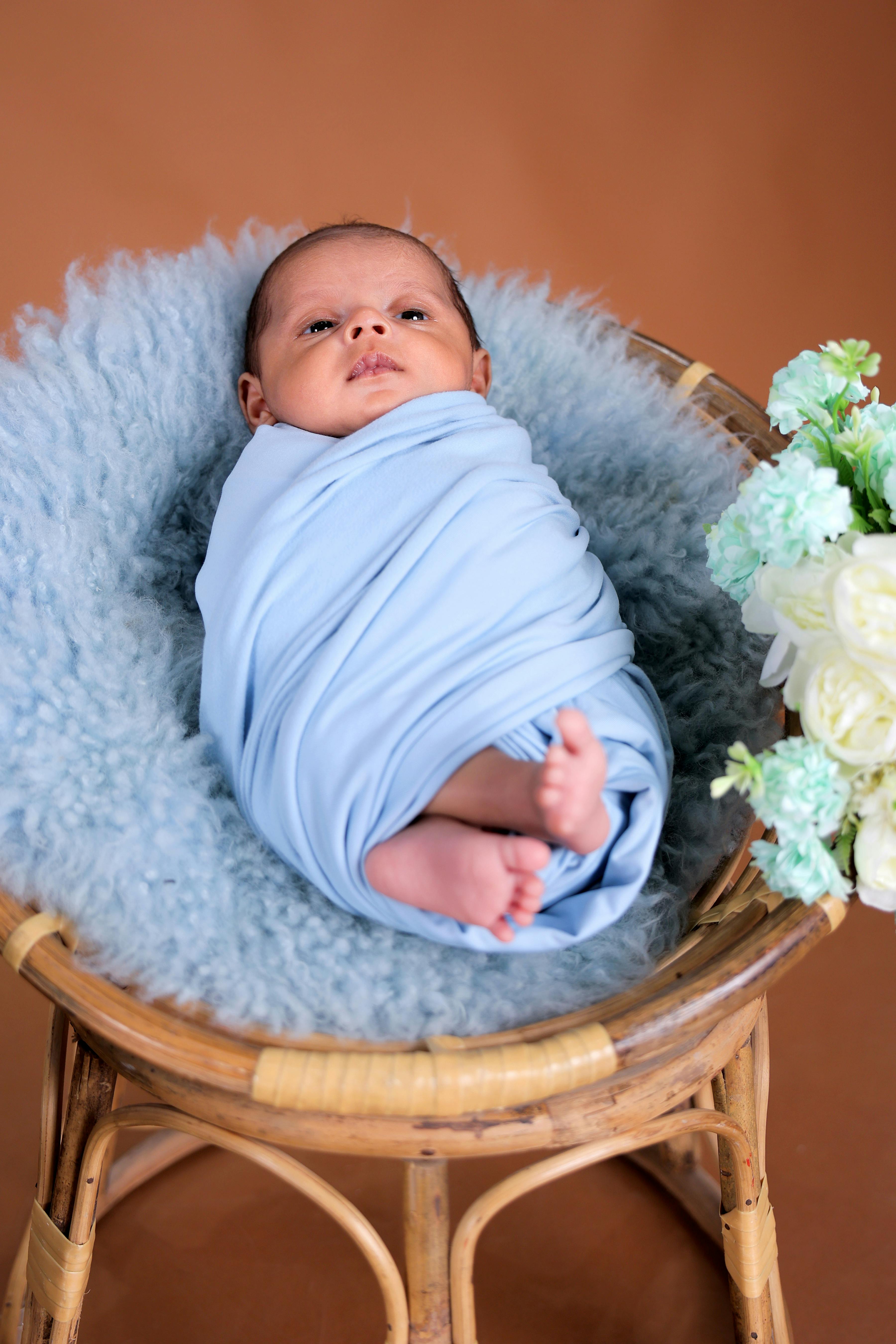 Adorable newborn baby in a blue wrap on a cozy chair with flowers, perfect for baby lifestyle themes.