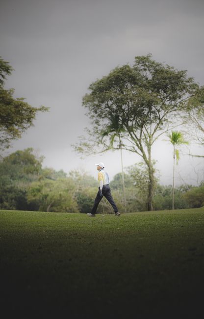 Fotomural Un golfista camina por un campo de golf rodeado de exuberante vegetación y árboles altos bajo un cielo nublado.