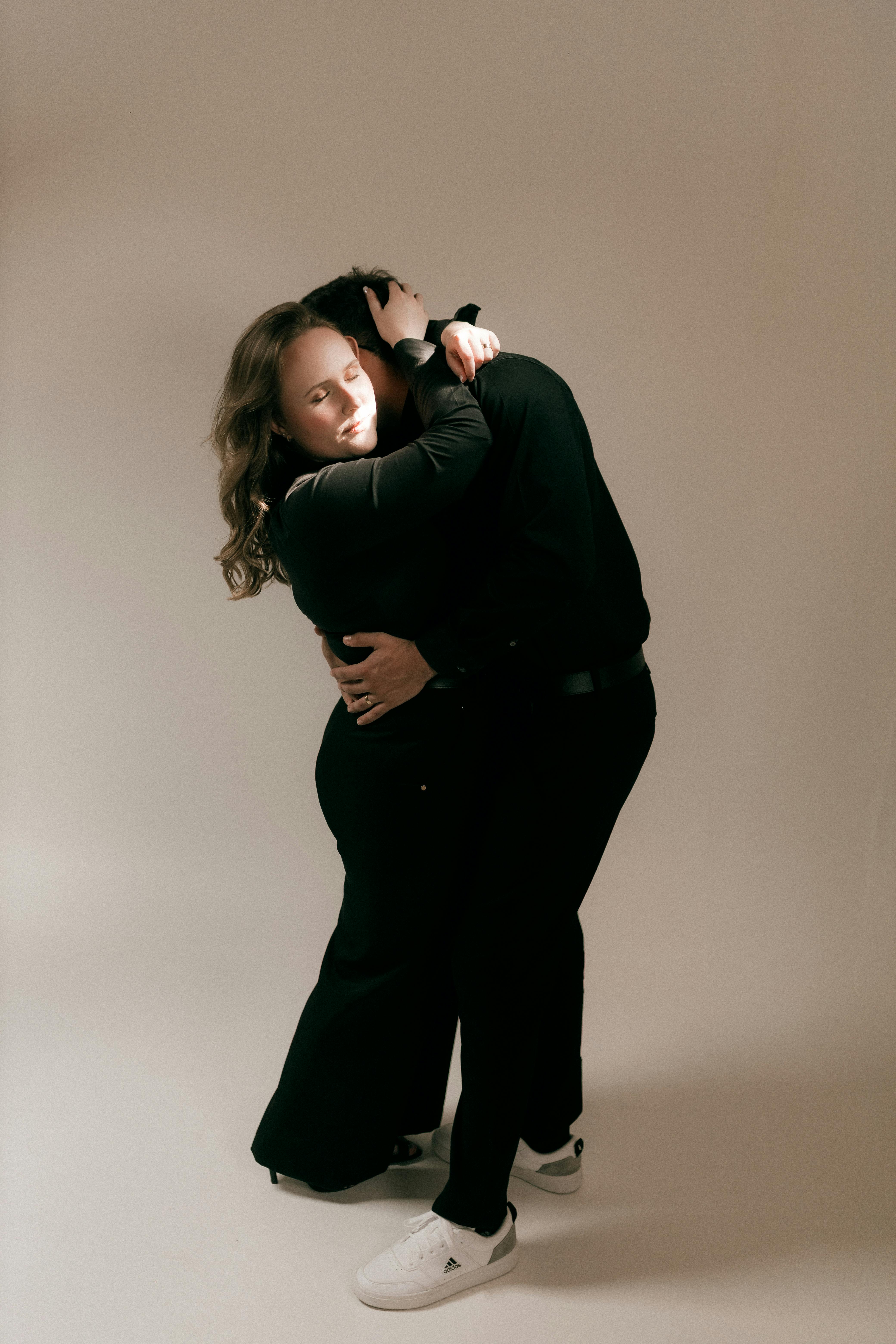 Gratis Una coppia di innamorati vestiti di nero si abbracciano intimamente sotto la calda luce dello studio. Foto a disposizione