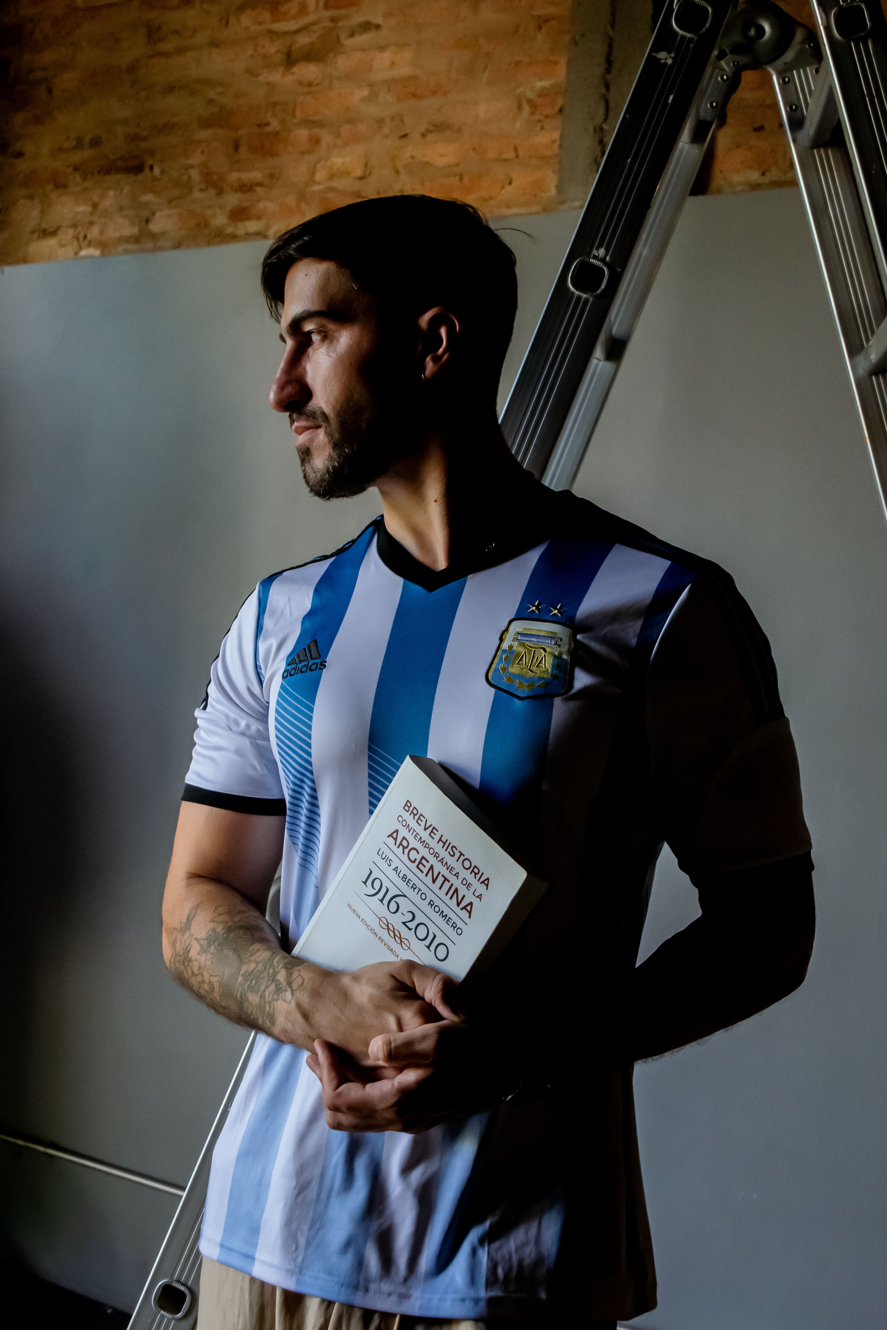 Man holding a history book wearing Argentina football jersey indoors.