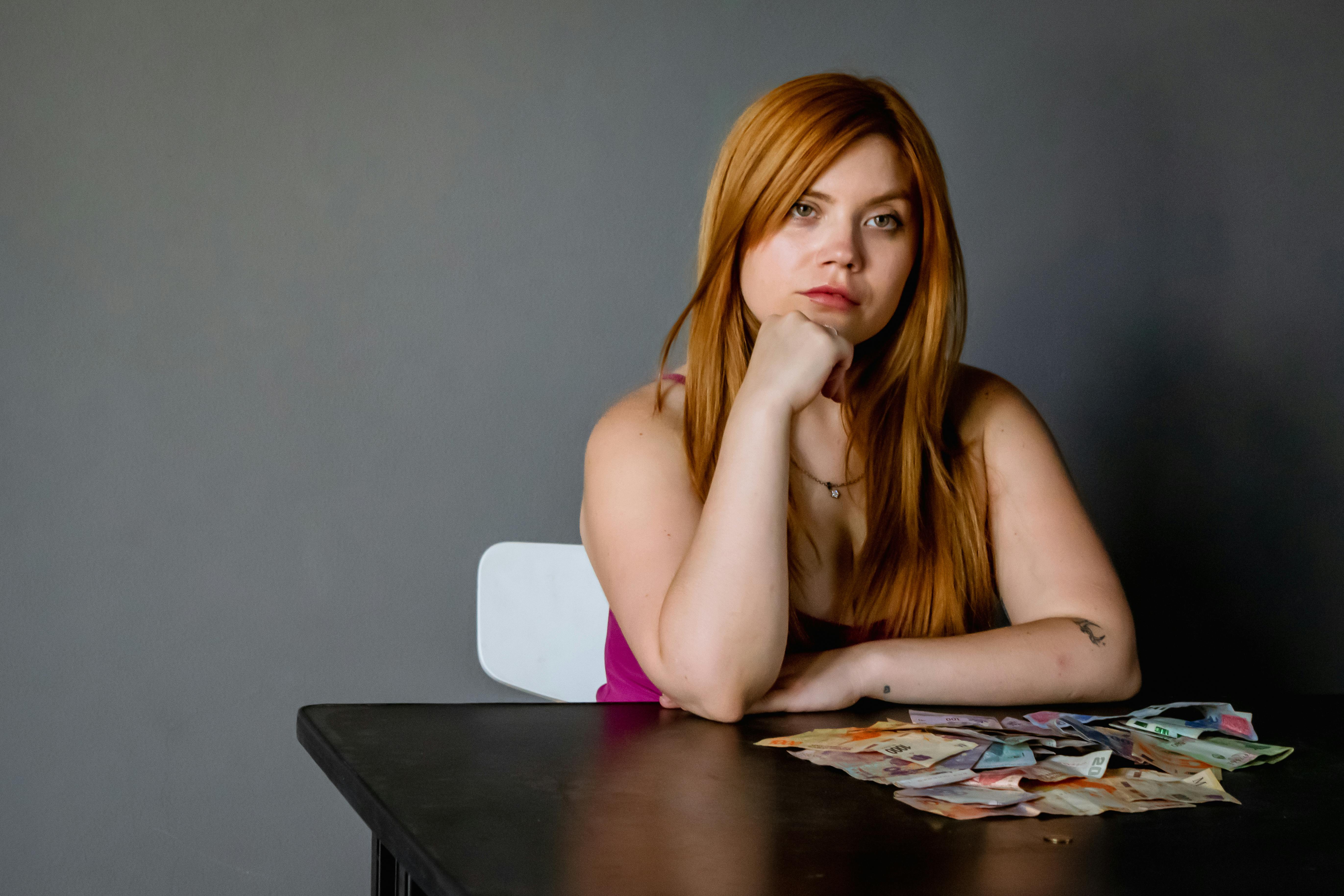 Young woman sitting at a table with assorted currency, looking contemplative.