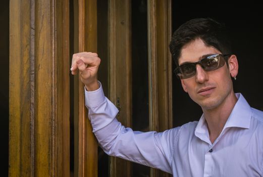 Confident man in sunglasses leaning on wooden door frame, wearing a white shirt.