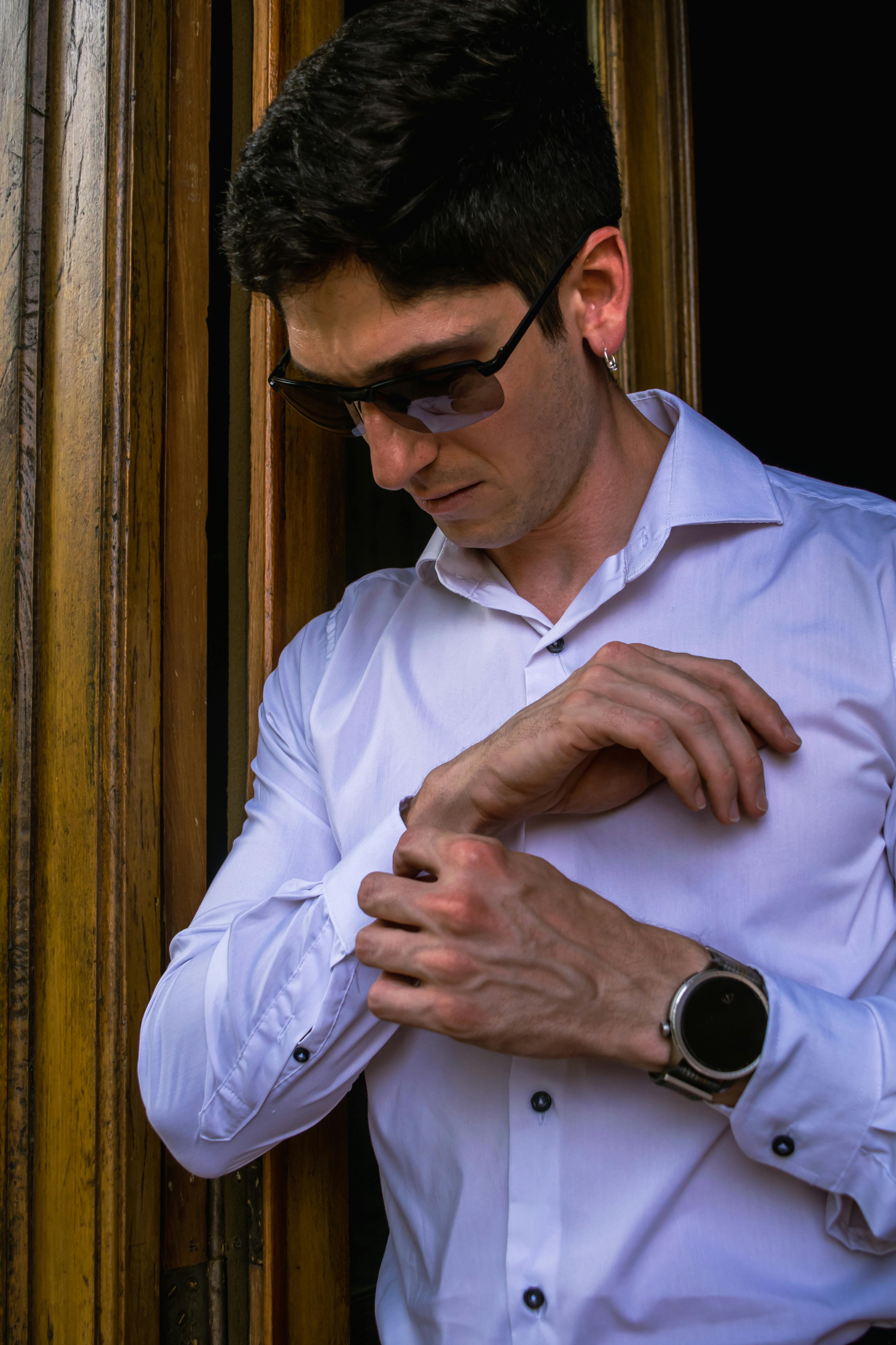 Free Man in white shirt adjusting cufflinks while standing near a wooden door, exuding style and elegance. Stock Photo