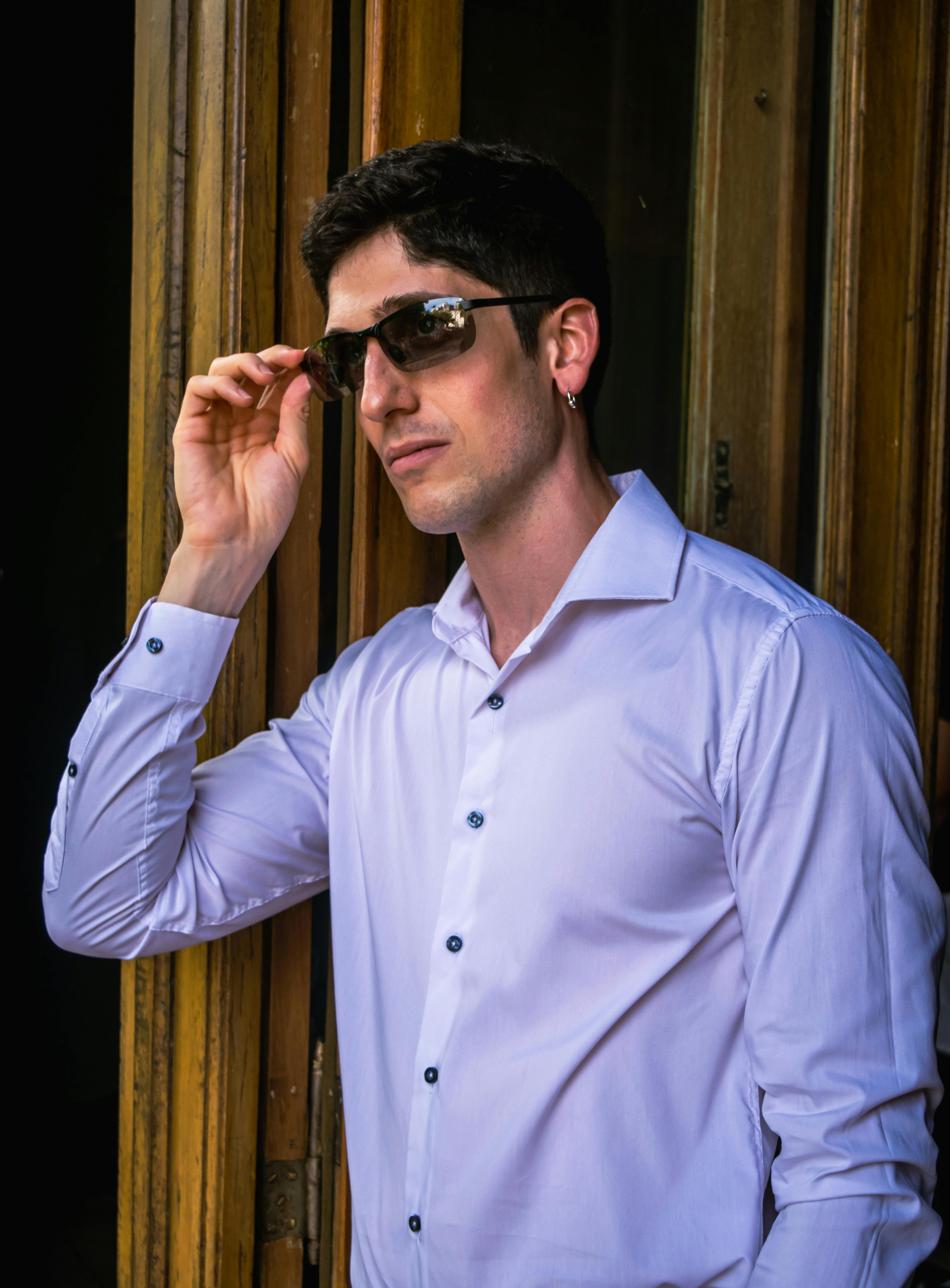 bezplatná Módní dospělý muž v slunečních brýlích a bílé košili, stojící u dřevěných dveří. Základová fotografie