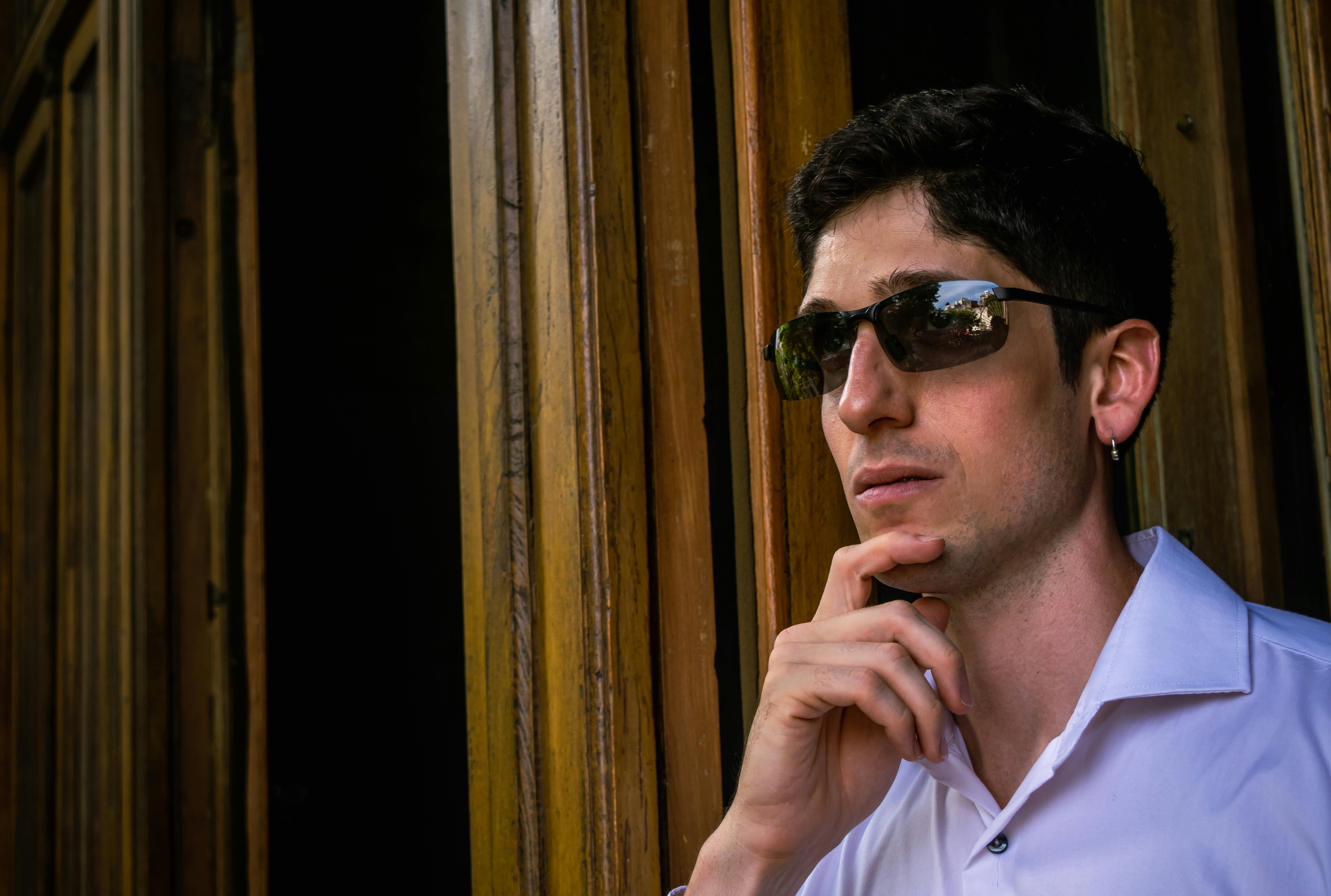 Gratis Un uomo pensieroso, con gli occhiali da sole, è in piedi accanto a pannelli di legno, immerso nella contemplazione. Foto a disposizione