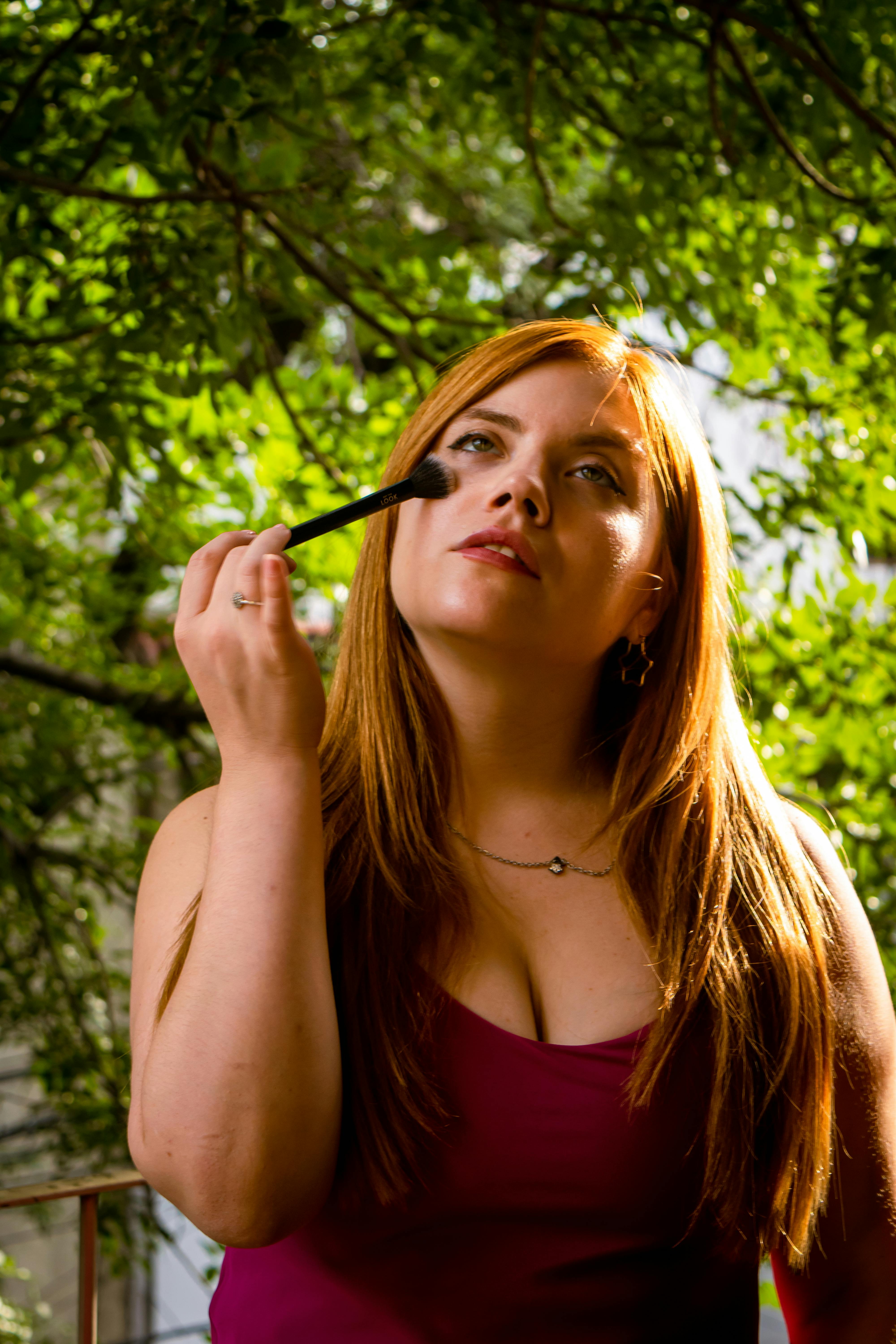 gratis Roodharige vrouw brengt make-up aan met een kwast in een zonnige buitenomgeving. Stockfoto