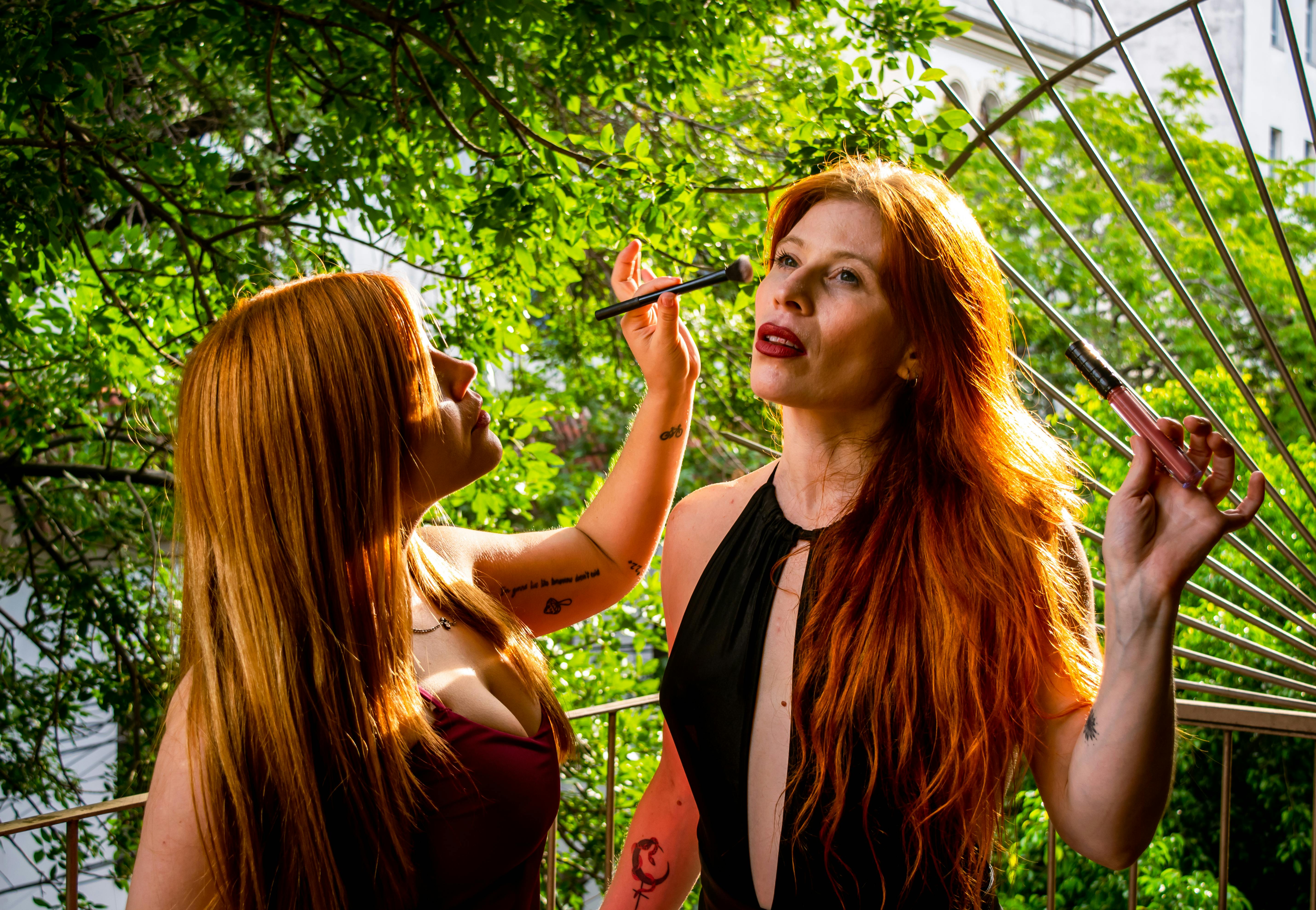 Ücretsiz Kızıl saçlı iki kadın, yemyeşil bitki örtüsüyle çevrili açık havada fırçalar kullanarak makyaj yapıyor. Stok Fotoğraflar